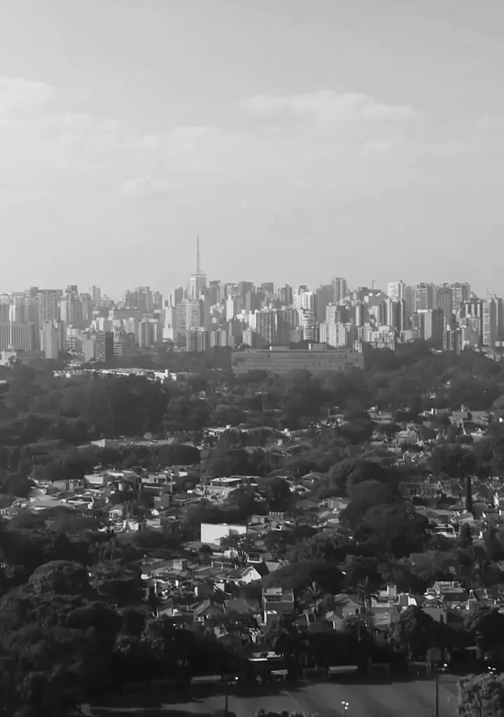 O entorno visto do alto, em preto e branco: as quadras e o campo do Clube Monte Líbano embaixo, o bairro de casas entre árvores e o skyline da cidade no horizonte.