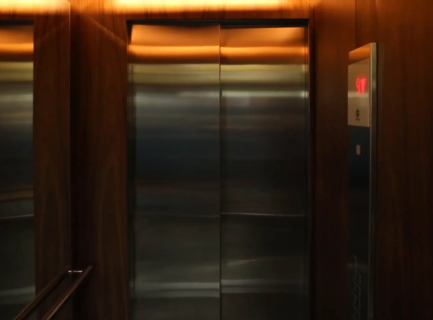 O hall dos elevadores: as portas de aço escovado entre painéis de madeira, com a luz embutida no forro.