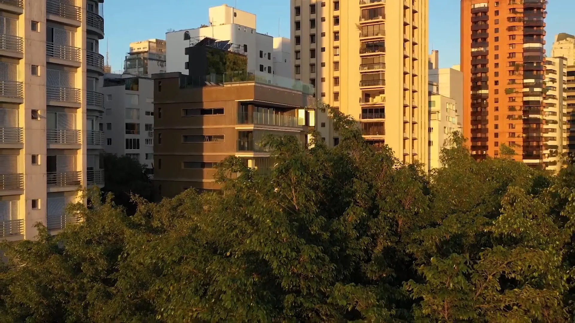 O Âmbar Jardins ao entardecer, entre as torres do bairro: a luz dourada nas fachadas, o rooftop com plantas no alto e as copas das árvores na frente.