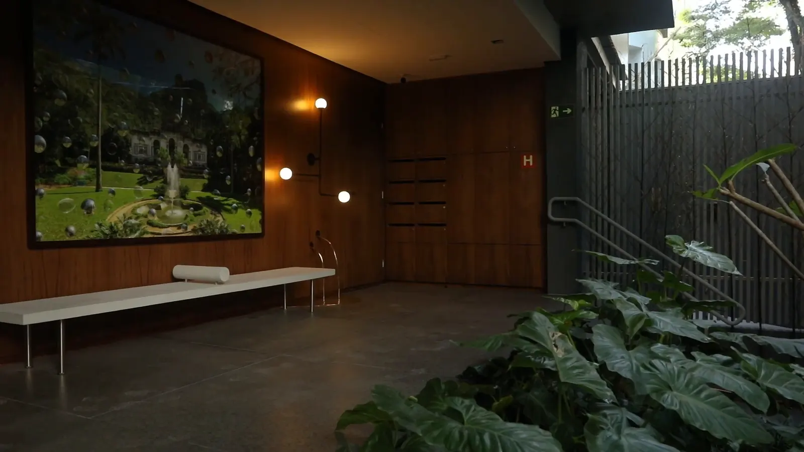 O hall social: as paredes de madeira escura, um quadro grande de um jardim, as arandelas acesas, um banco branco comprido e folhagens junto ao muro de ripas.