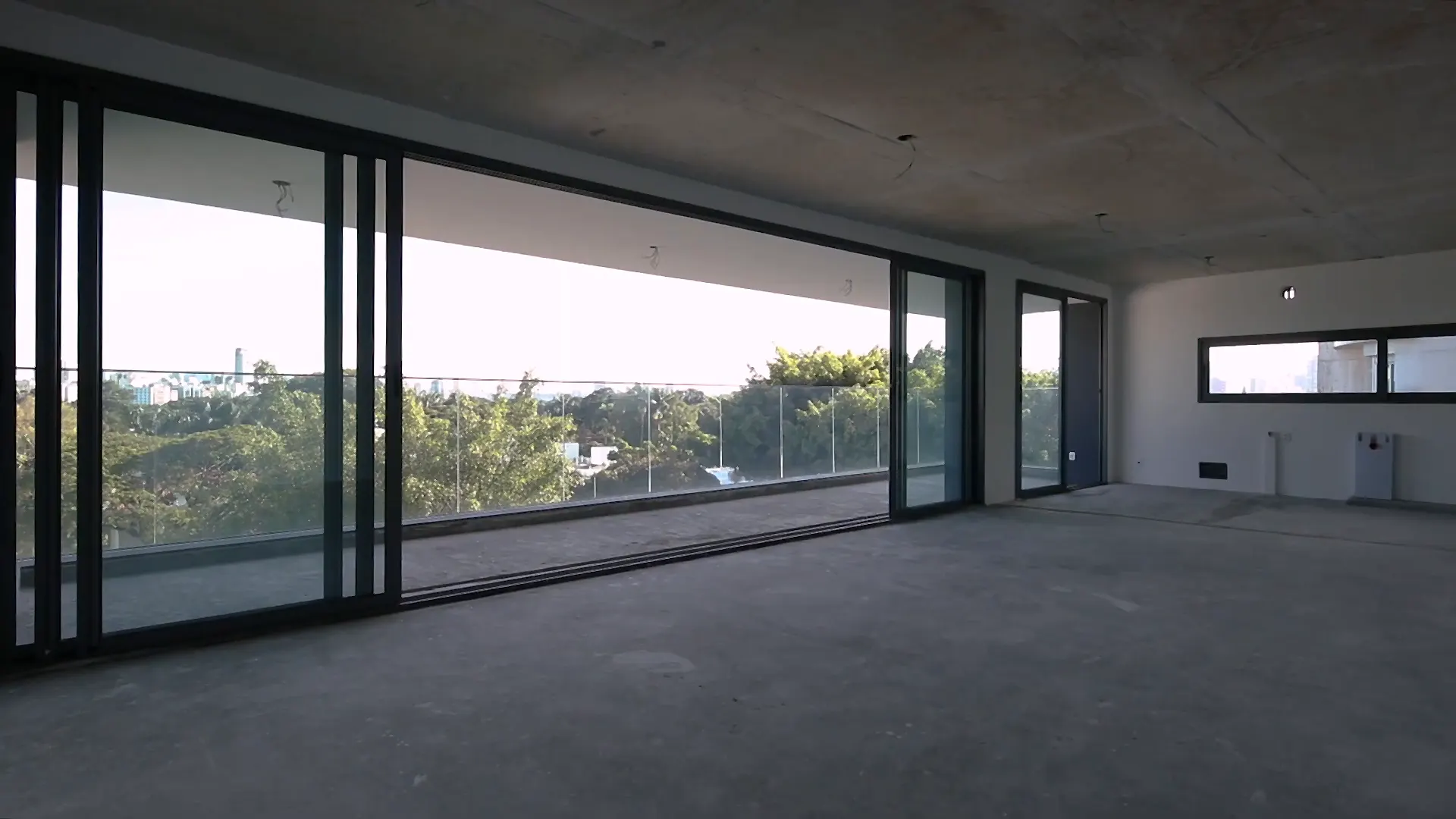 O living de uma unidade entregue, ainda sem piso: os caixilhos de correr do chão ao teto abertos para a varanda de vidro e, lá fora, as copas das árvores e os prédios ao longe.