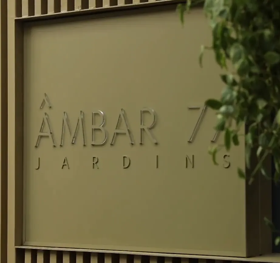A placa da entrada de perto: o painel cáqui emoldurado pelas ripas, com as letras de aço ÂMBAR 77 e, embaixo, JARDINS espaçado, e uma trepadeira à direita.