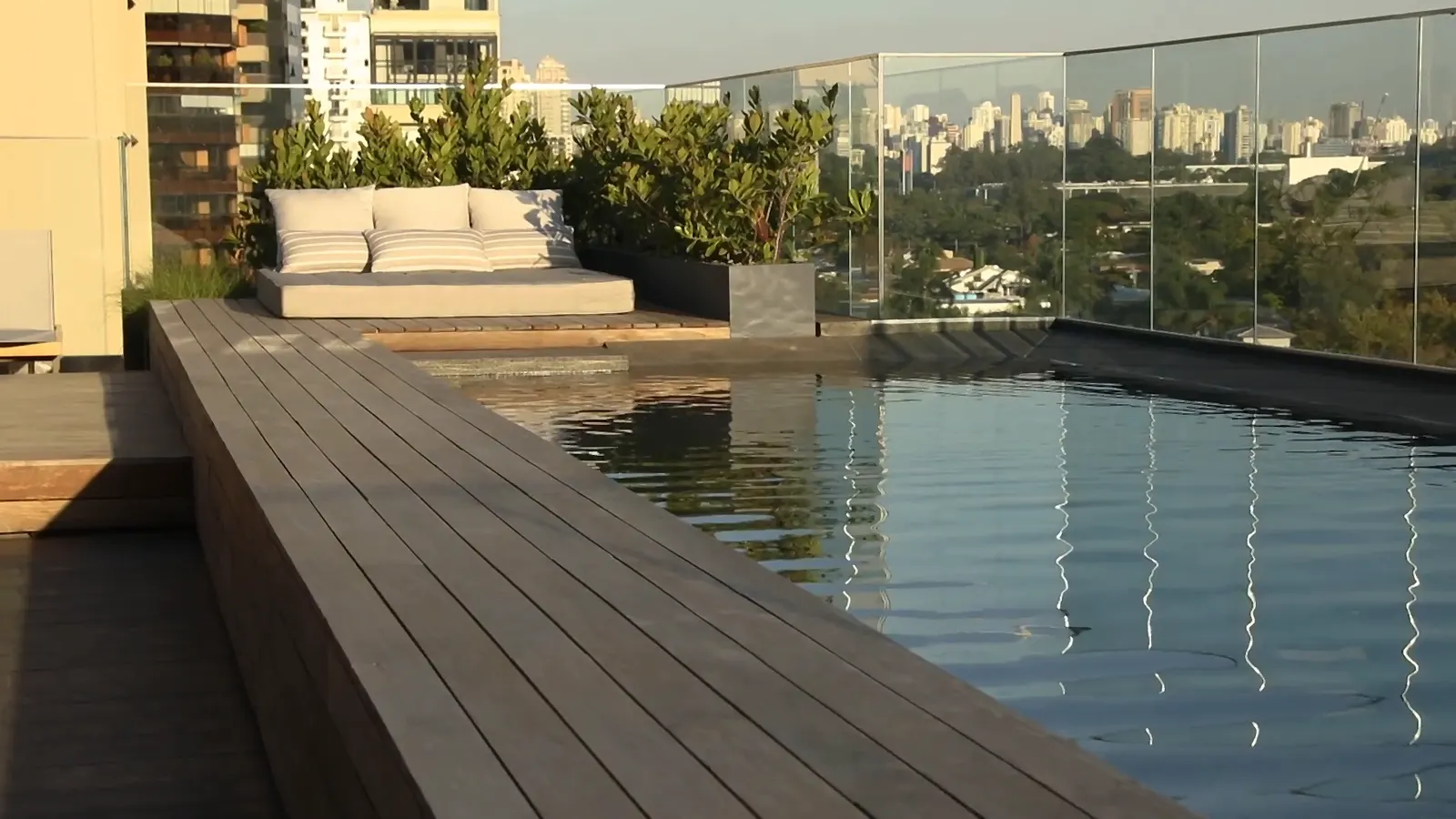 O deque de madeira ao lado da piscina, o futon com almofadas listradas, as plantas em jardineiras e o guarda-corpo de vidro, com a cidade ao fundo.