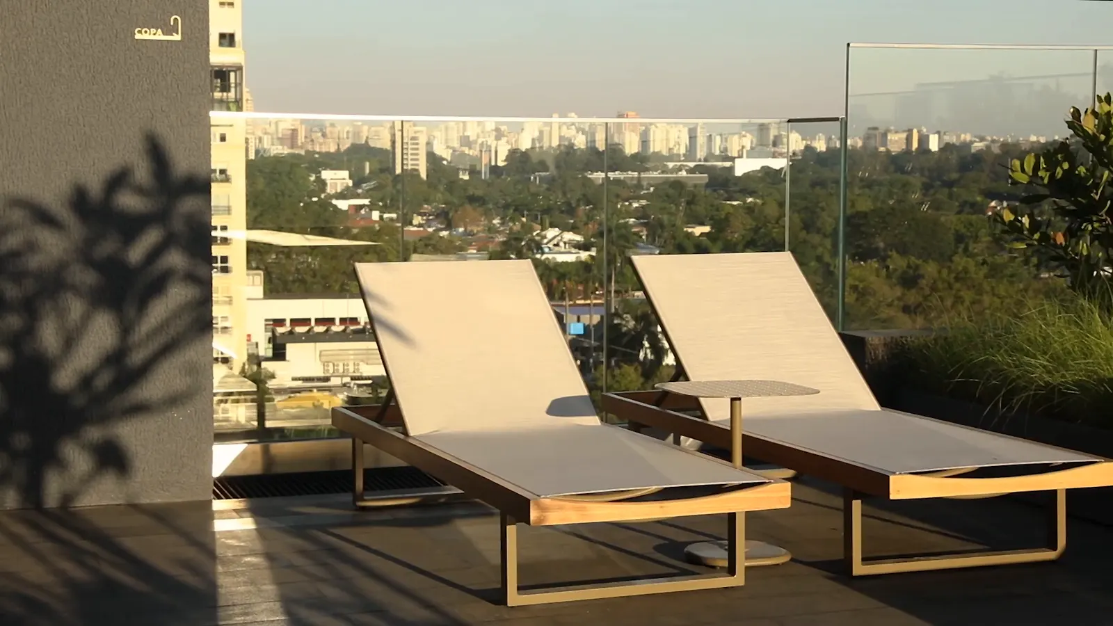 Duas espreguiçadeiras claras sobre o deque do rooftop, à luz do fim da tarde, com a sombra das plantas e o horizonte de prédios.