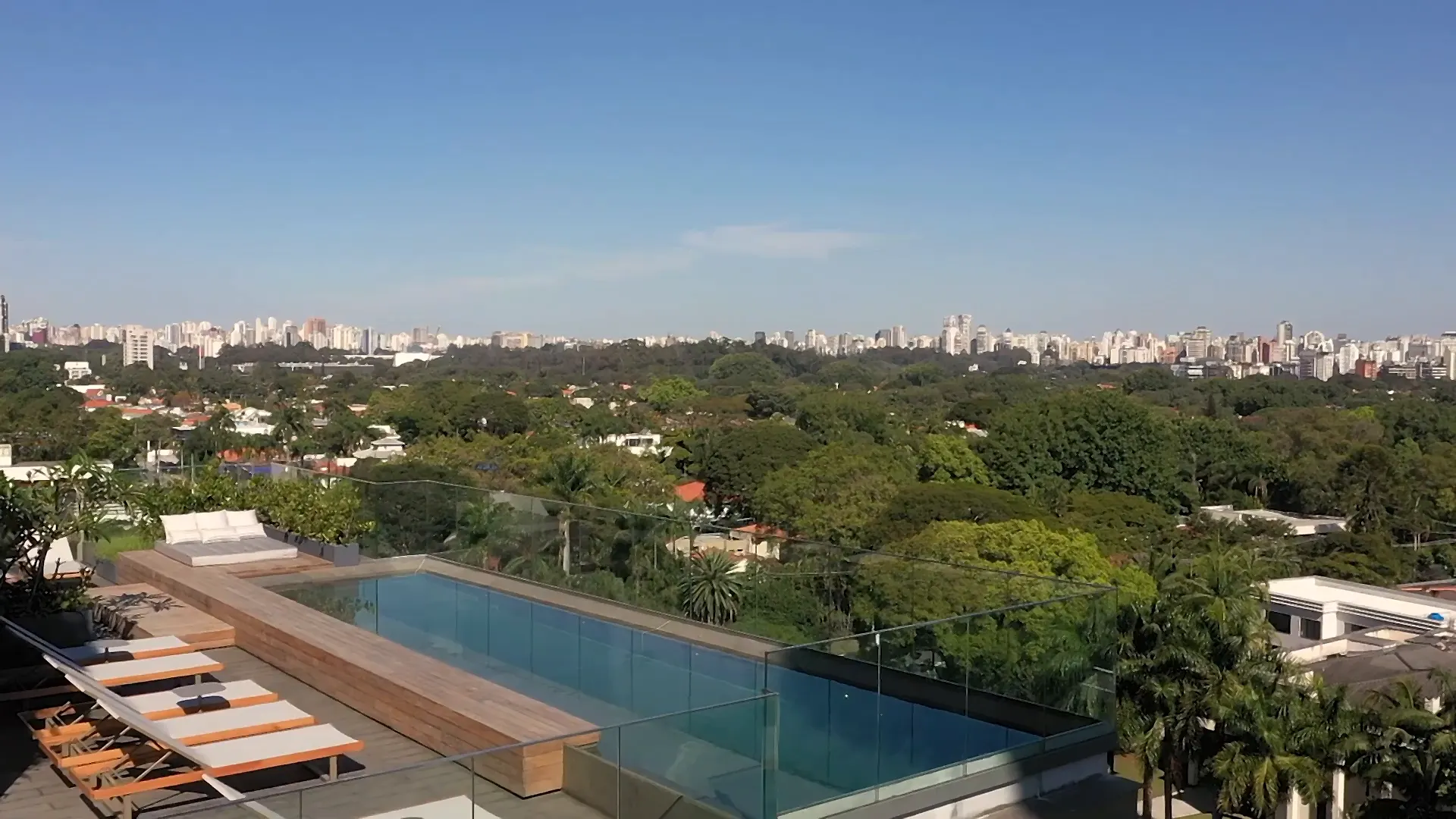 O rooftop pronto, de cima: a piscina de borda infinita com o deque de madeira e as espreguiçadeiras, o futon com almofadas e, à frente, o verde do Jardim América até os prédios no horizonte.