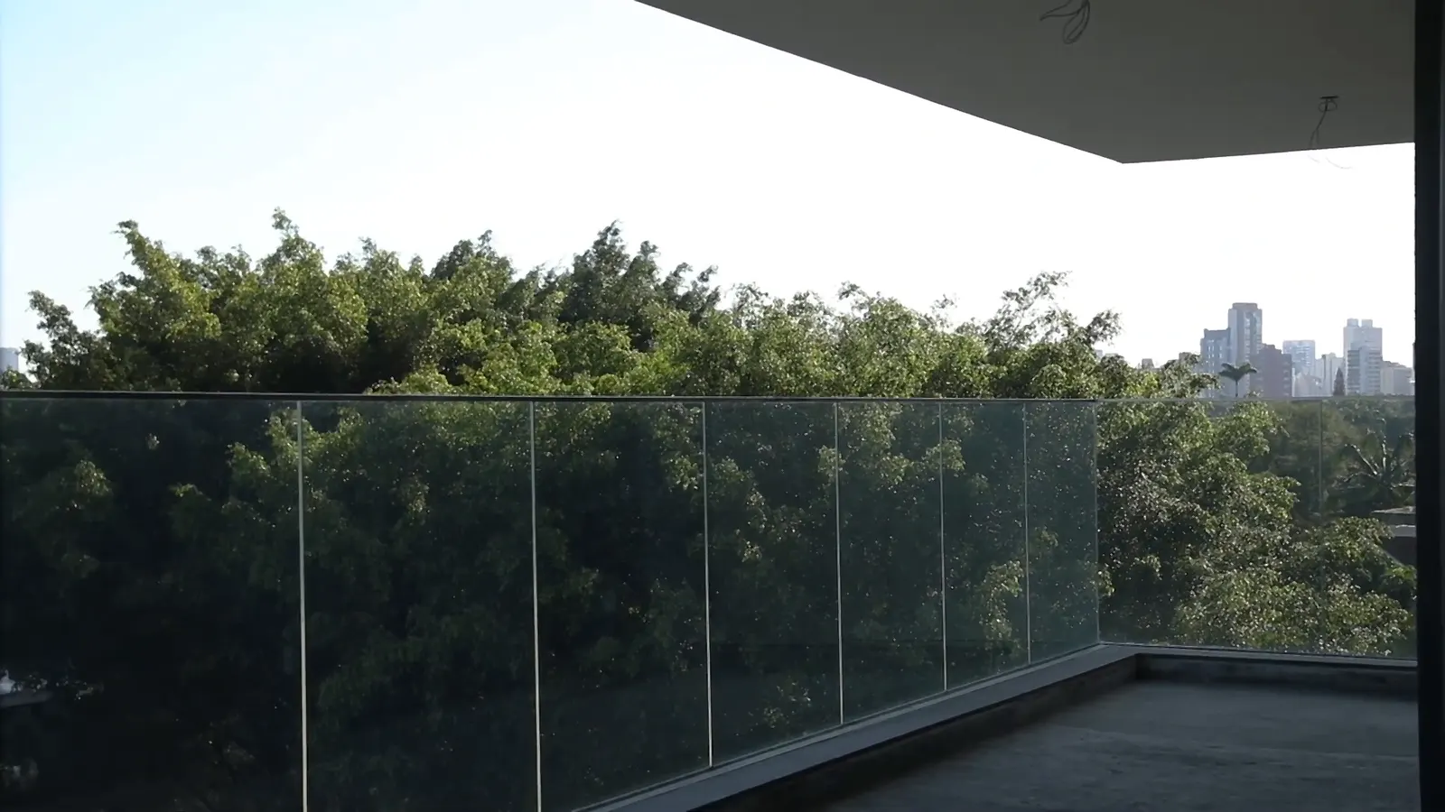 A varanda de uma unidade entregue: o guarda-corpo de vidro, a laje de cima em balanço e, na frente, uma mata alta de bambus e árvores, com prédios no horizonte.