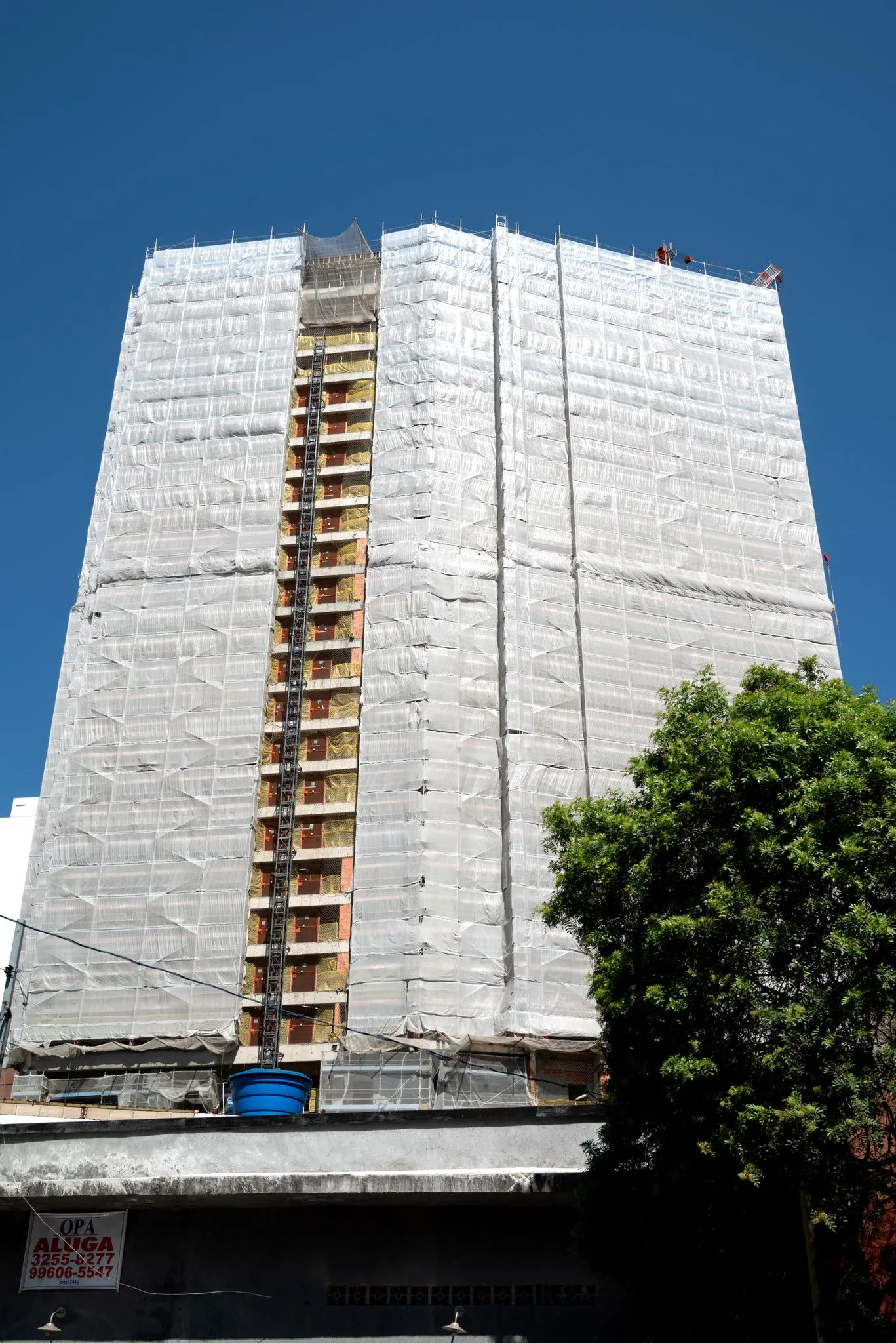 Fotografia da obra: a torre em estrutura, coberta por telas de proteção, contra o céu azul, com uma árvore na calçada.