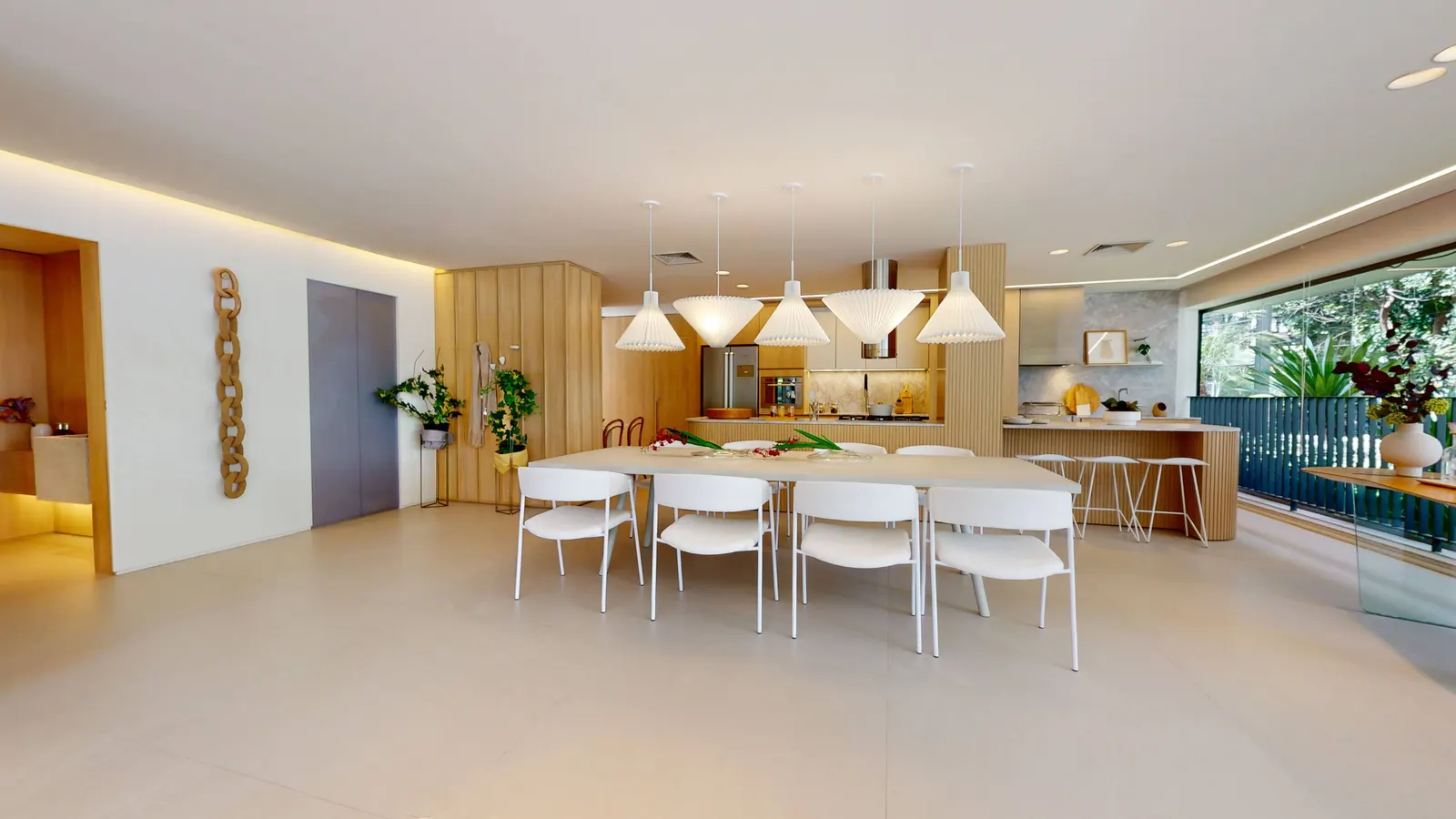 A mesa de jantar longa com cadeiras brancas sob luminárias pendentes, a cozinha de madeira ao fundo e o terraço à direita.