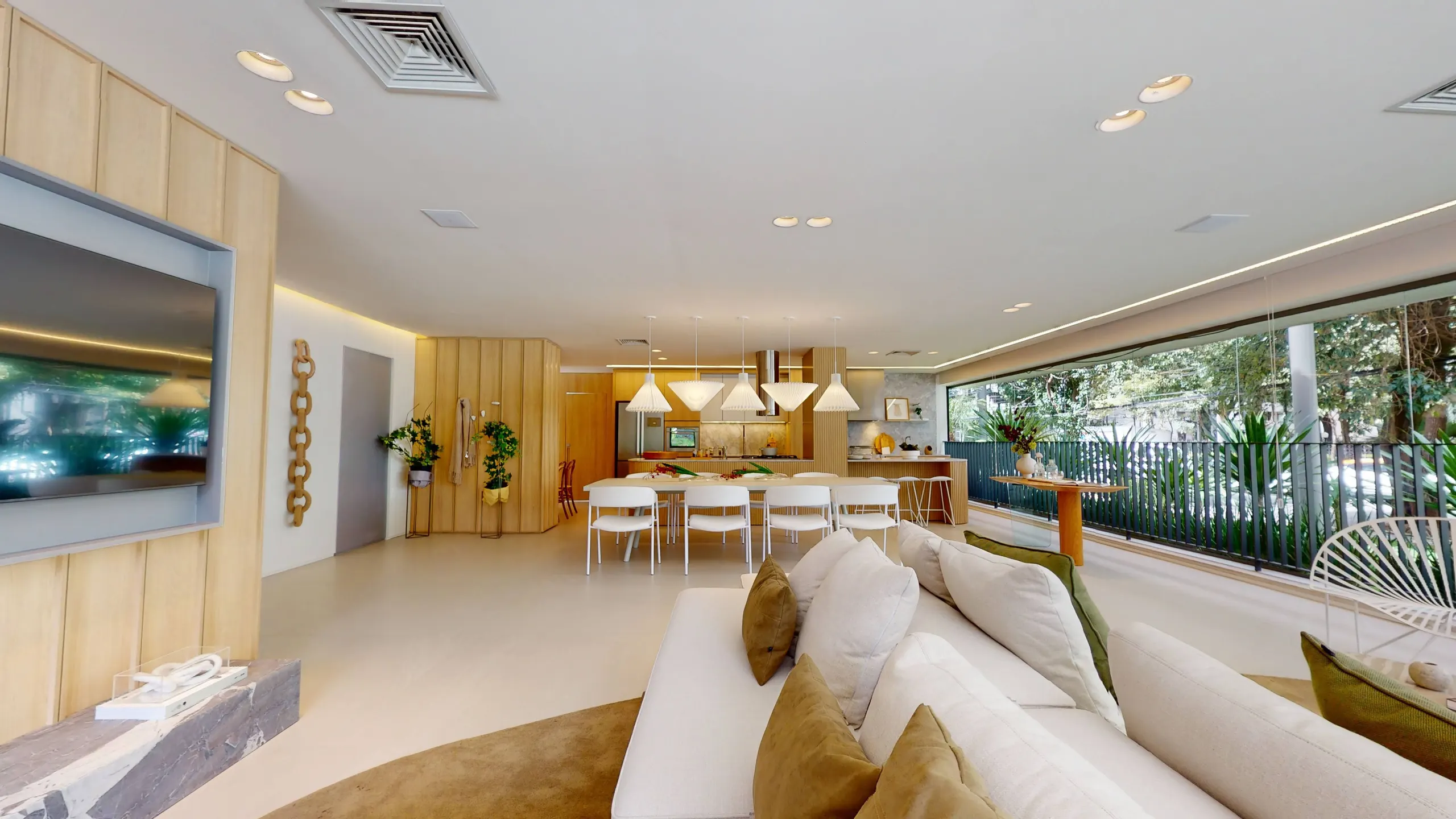 O living do decorado: o sofá claro diante do painel de madeira com a TV, e ao fundo a mesa de jantar e a parede de vidro aberta para o verde.