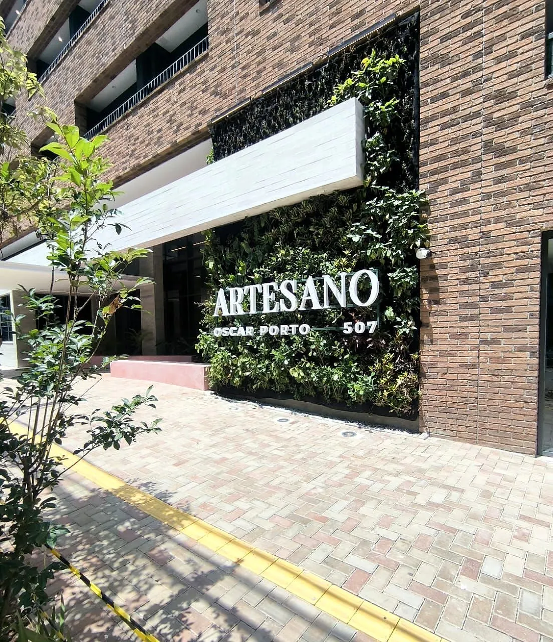 A entrada do residencial: o letreiro branco ARTESANO OSCAR PORTO 507 sobre o jardim vertical, ao lado da parede de tijolo, com a calçada de pedra na frente.