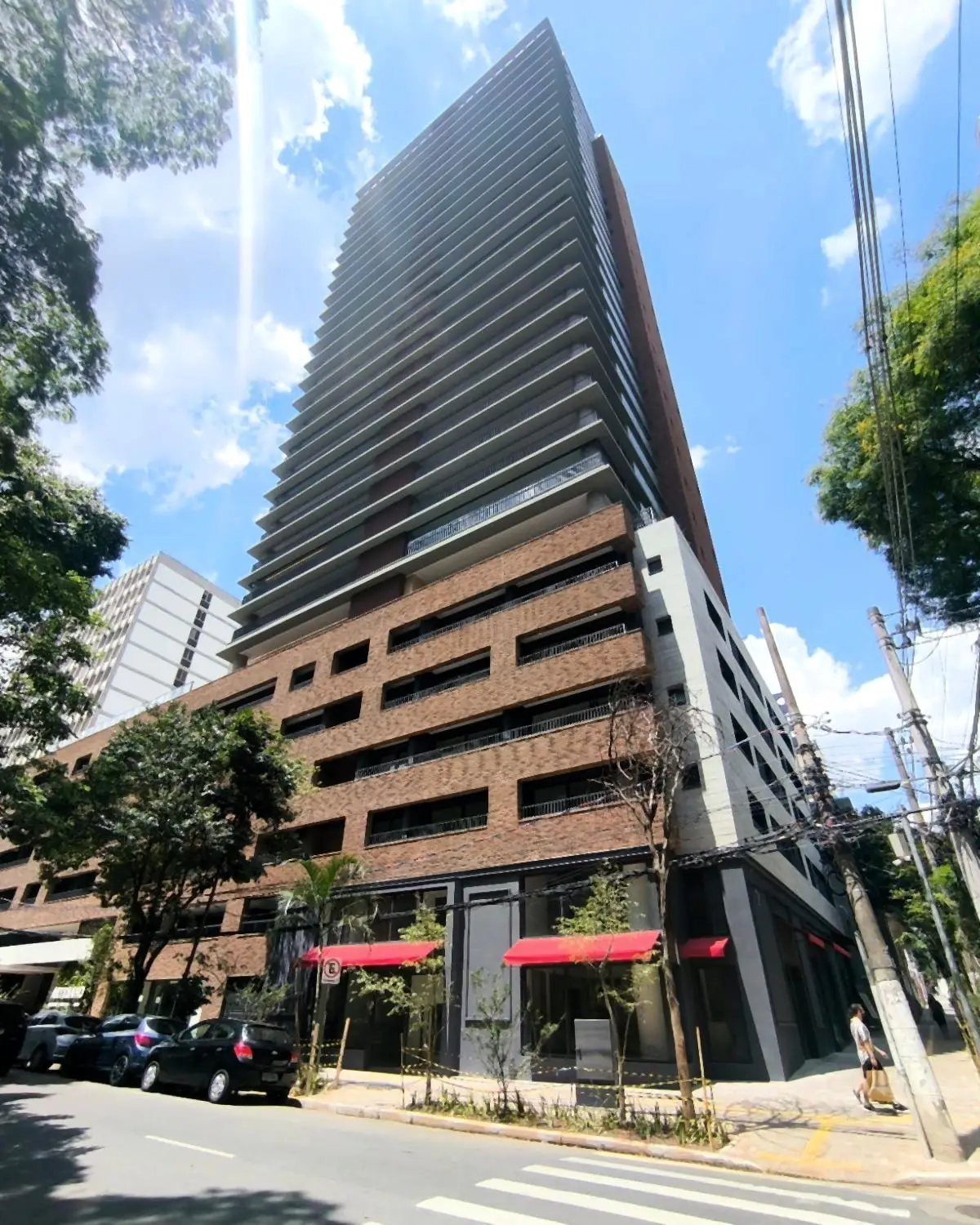 O Artesano Oscar Porto pronto, visto da esquina: a torre de varandas em linhas horizontais sobre a base de tijolo, com as lojas de toldo vermelho no térreo, árvores na calçada e o céu azul.