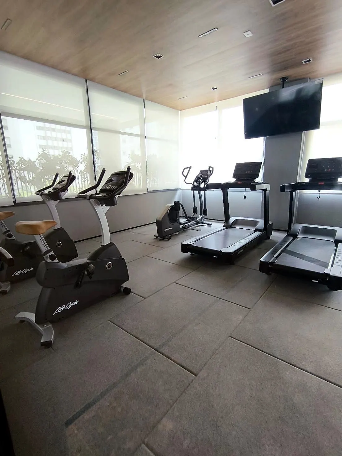 O fitness: bicicletas e esteiras sobre o piso de borracha, sob o forro de madeira, com a TV e as janelas de cortina clara.