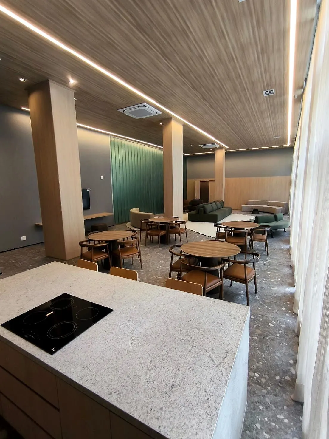 A área gourmet do salão: a bancada de pedra clara com o cooktop em primeiro plano e as mesas do salão atrás.
