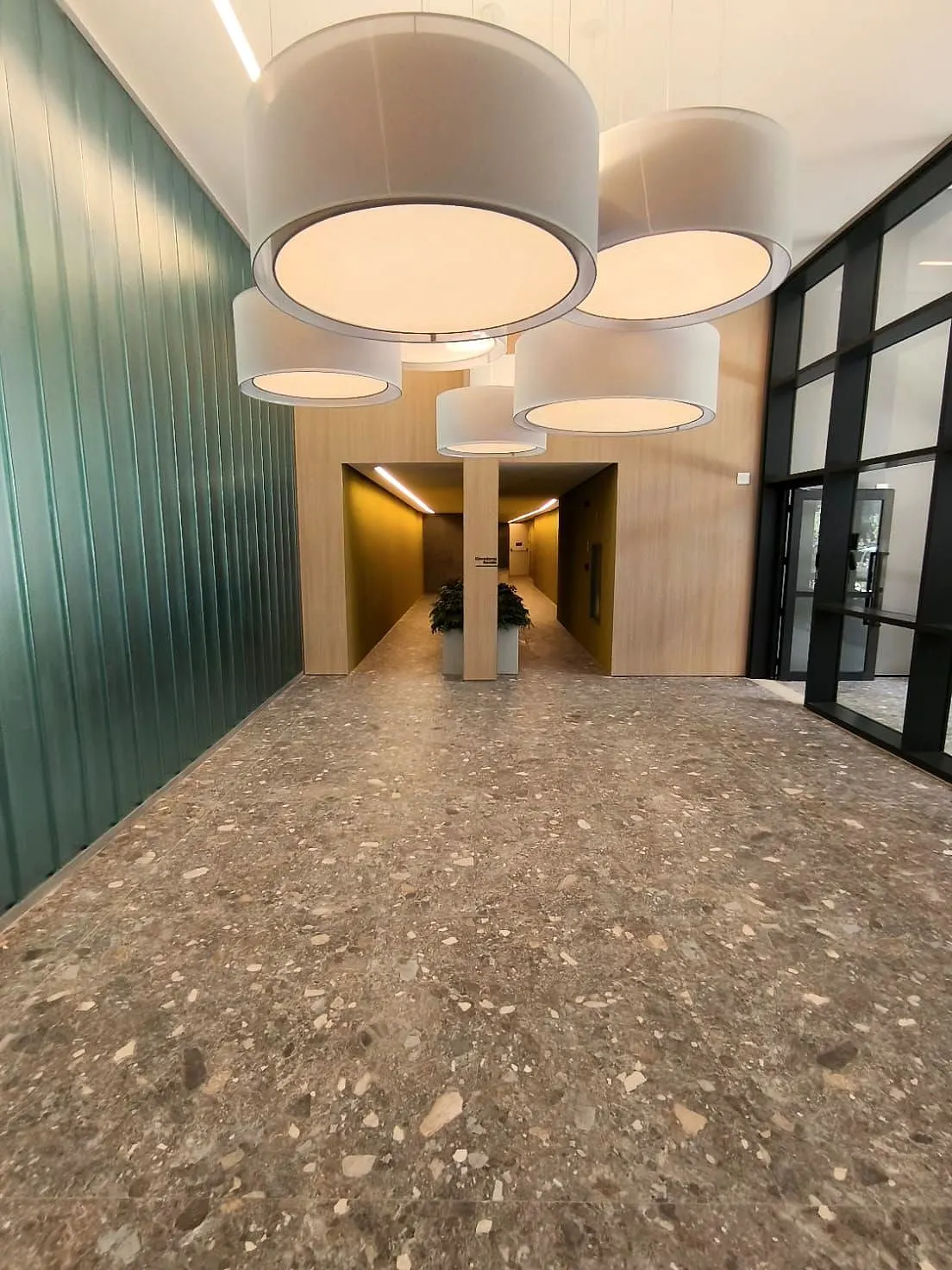 O lobby: o piso de granilite, a parede de painéis verdes, as luminárias brancas em cilindro penduradas e o corredor dos elevadores ao fundo.