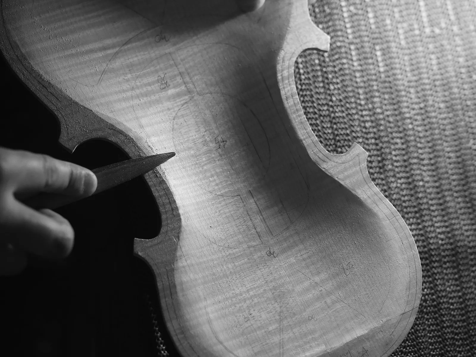 Em preto e branco, a mão de um luthier entalha a borda curva do tampo de um violino.