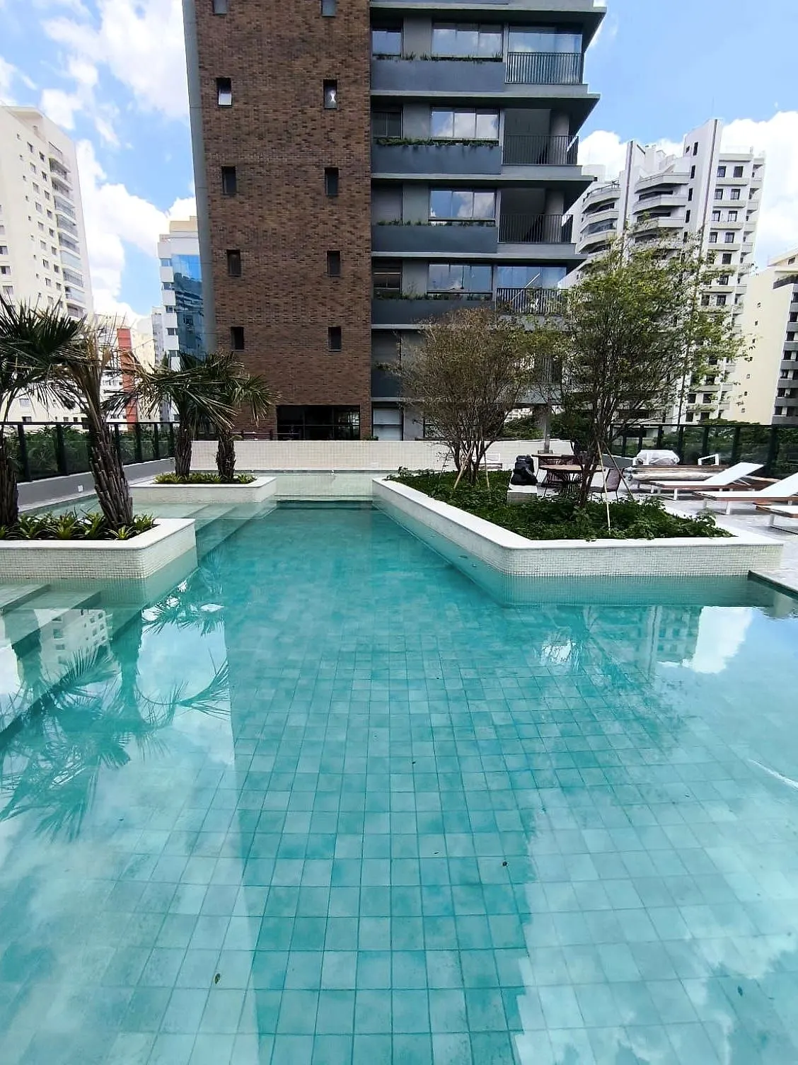 A piscina do lazer elevado: a água azul de pastilhas, os canteiros com árvores no meio e a torre de tijolo e vidro ao fundo.