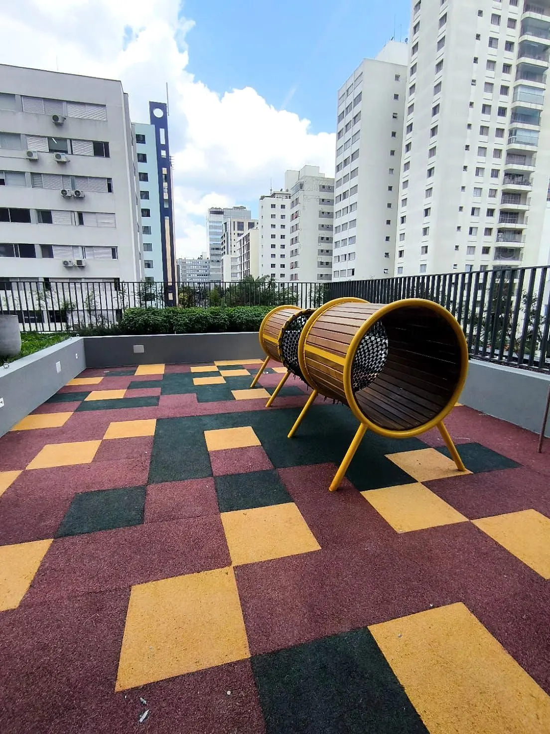 O playground ao ar livre: o piso de borracha em quadrados vermelhos, amarelos e verdes, com um túnel de madeira.