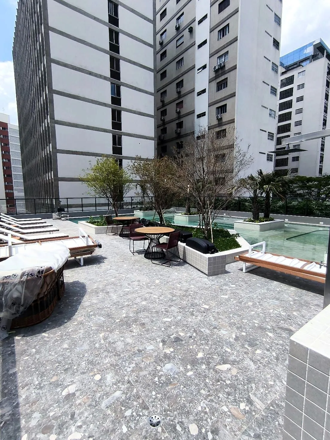O solarium: o deque de pedra com espreguiçadeiras e mesas, árvores em canteiros e a piscina ao fundo, entre os prédios do bairro.