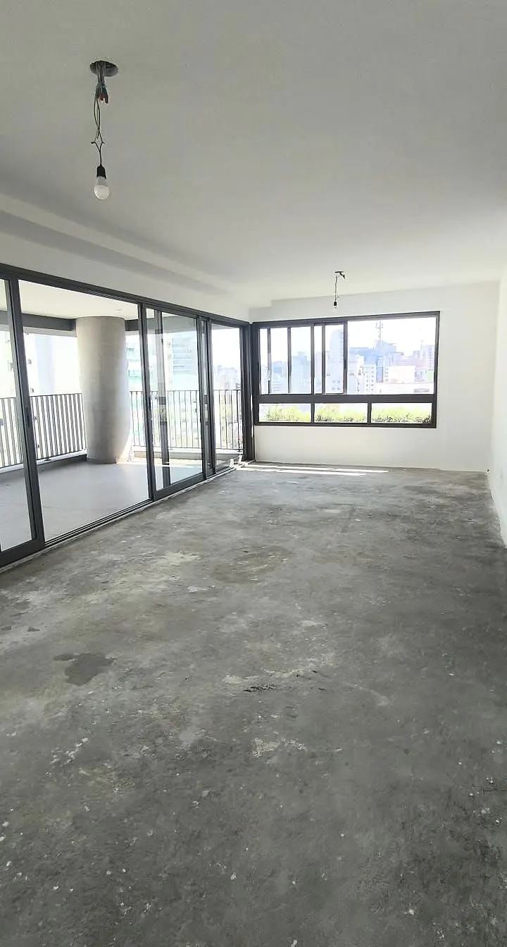 A sala da unidade 181 vazia, no contrapiso: o piso cinza de cimento, o teto branco com o fio da luz e as portas de vidro para o terraço, com prédios lá fora.