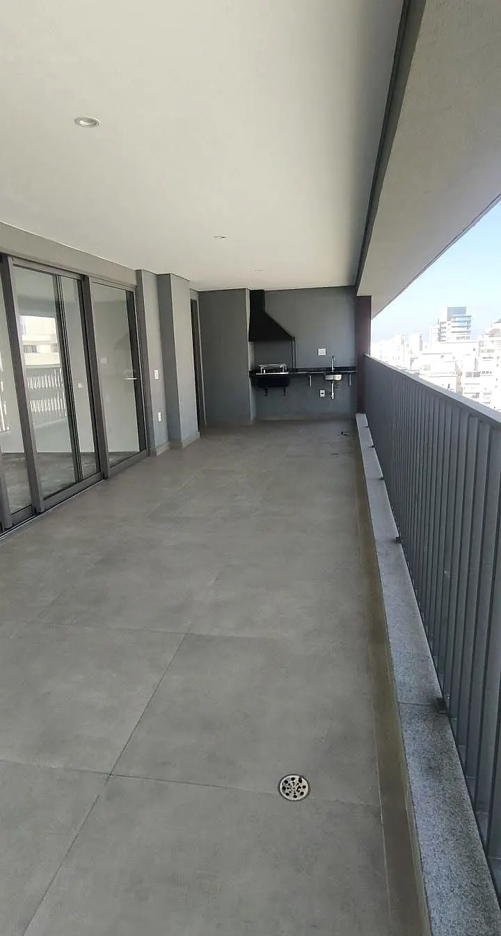 O terraço da unidade 181: o piso de porcelanato cinza, o guarda-corpo escuro à direita, as portas de vidro à esquerda e a churrasqueira no fundo.