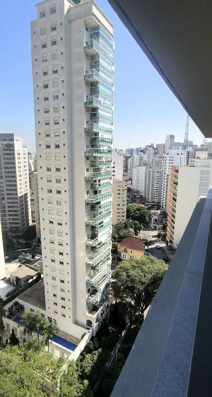 A vista do terraço da unidade 181, no 18º andar: uma torre branca alta ao lado, prédios do bairro e as copas das árvores lá embaixo.