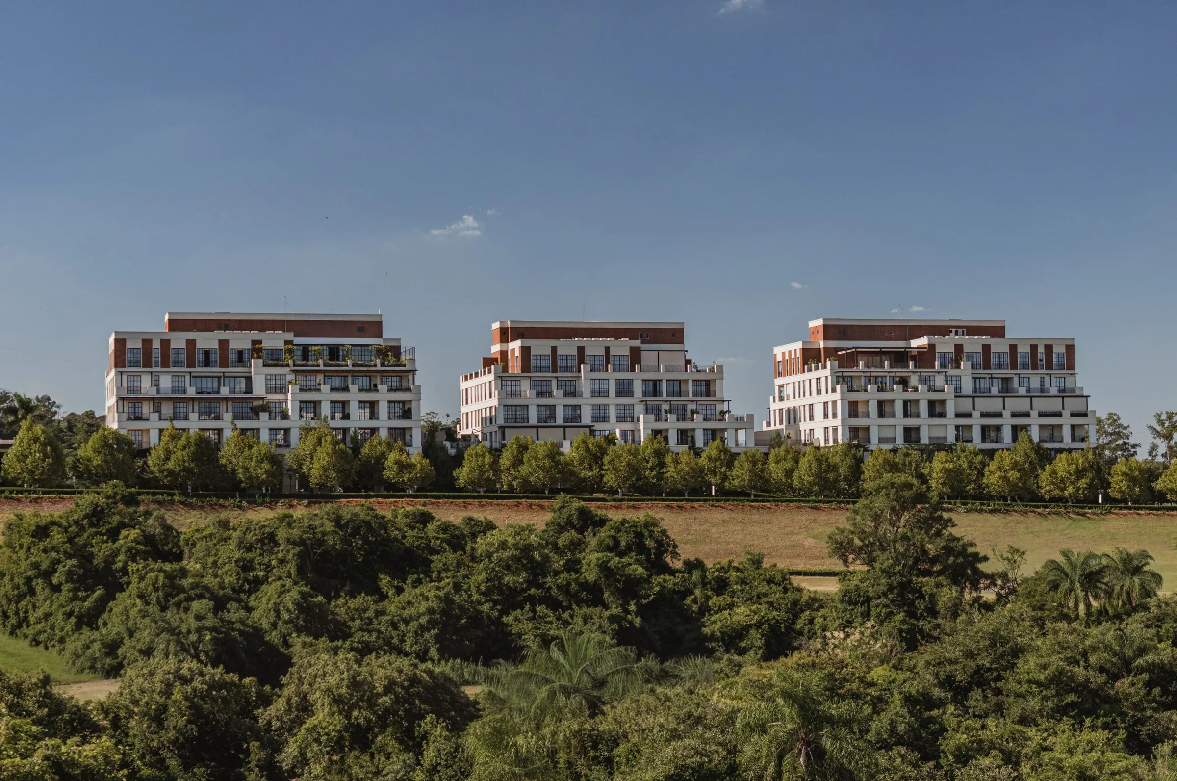 Os três edifícios das Golf Residences acima da mata, com o gramado do campo de golfe à frente, sob céu azul.