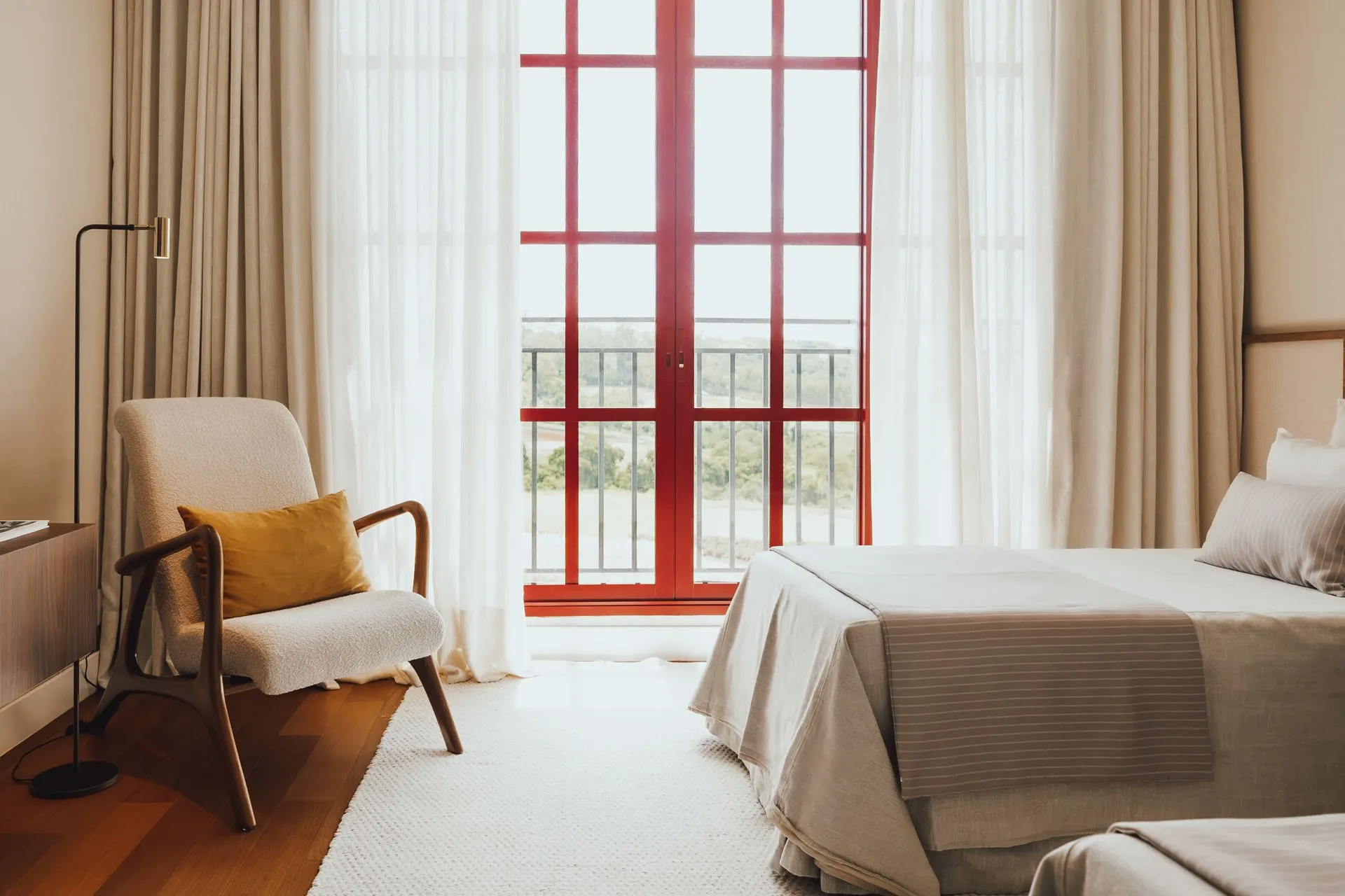 Suíte decorada de uma unidade das Golf Residences, com cama de casal, poltrona e janelas de caixilho vermelho do piso ao teto.