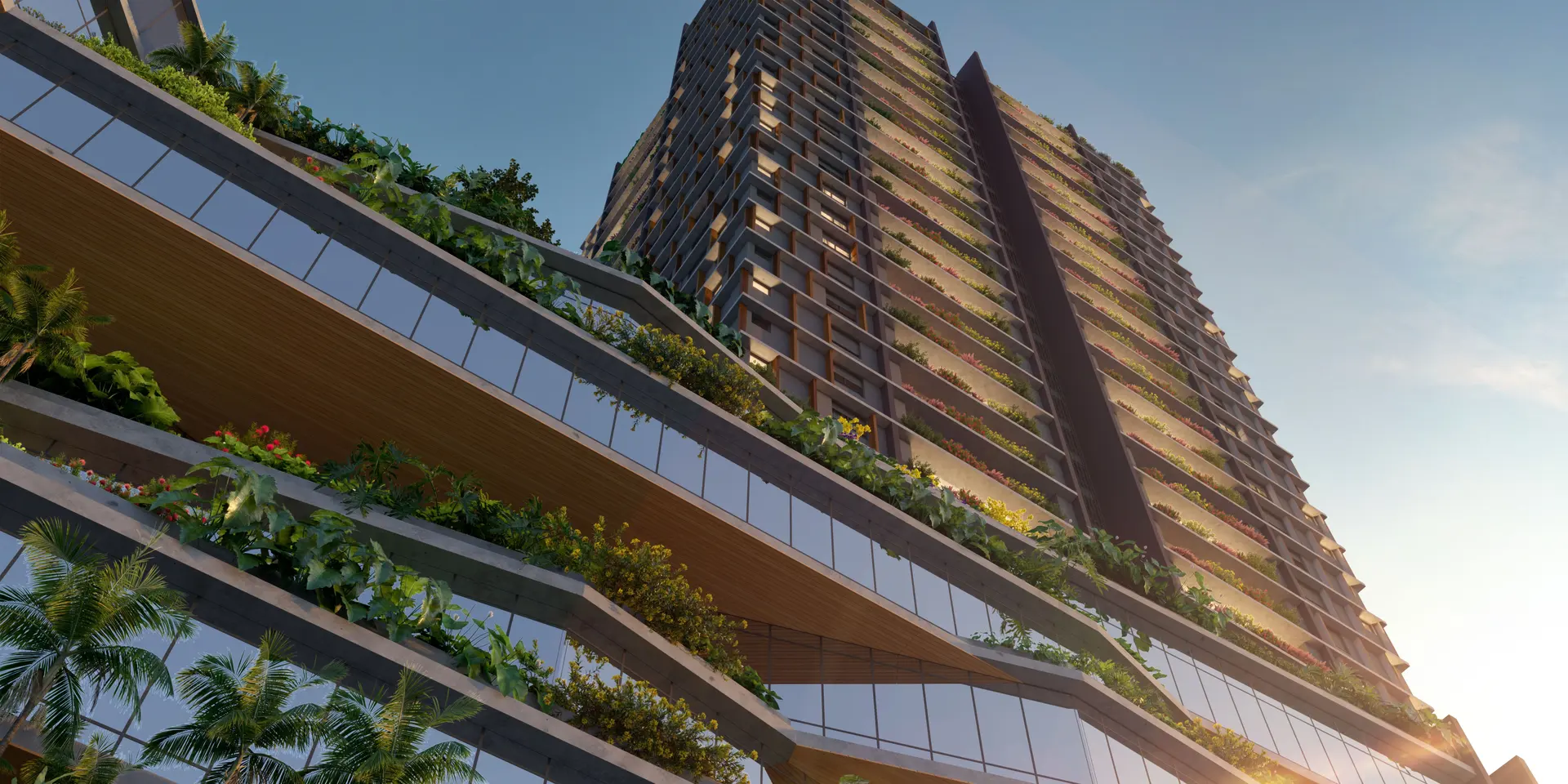 A fachada vista de baixo, ao entardecer: faixas inclinadas de jardineiras floridas e vidro sobem pela base, e a torre de terraços verdes se ergue atrás, contra o céu.