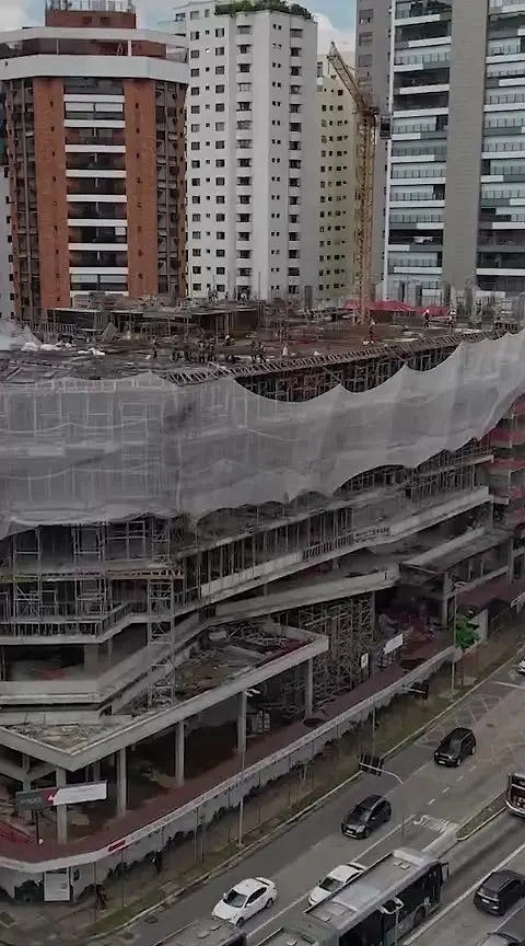 A obra vista do alto, em setembro de 2026: os andares de concreto com a tela de proteção, os andaimes, a avenida embaixo e os prédios do bairro em volta.