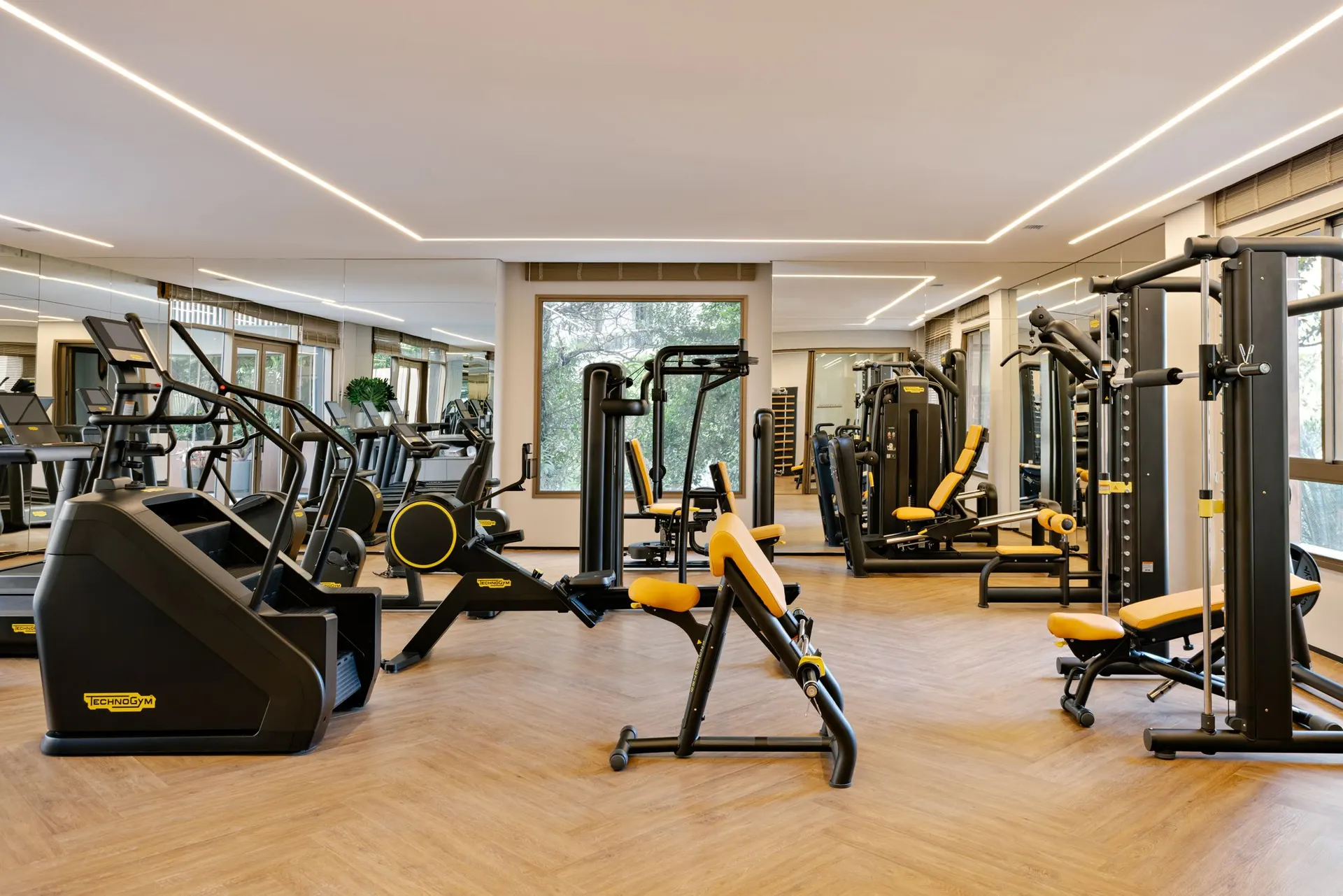 A academia com equipamentos Technogym, piso de madeira e janelas para o jardim.
