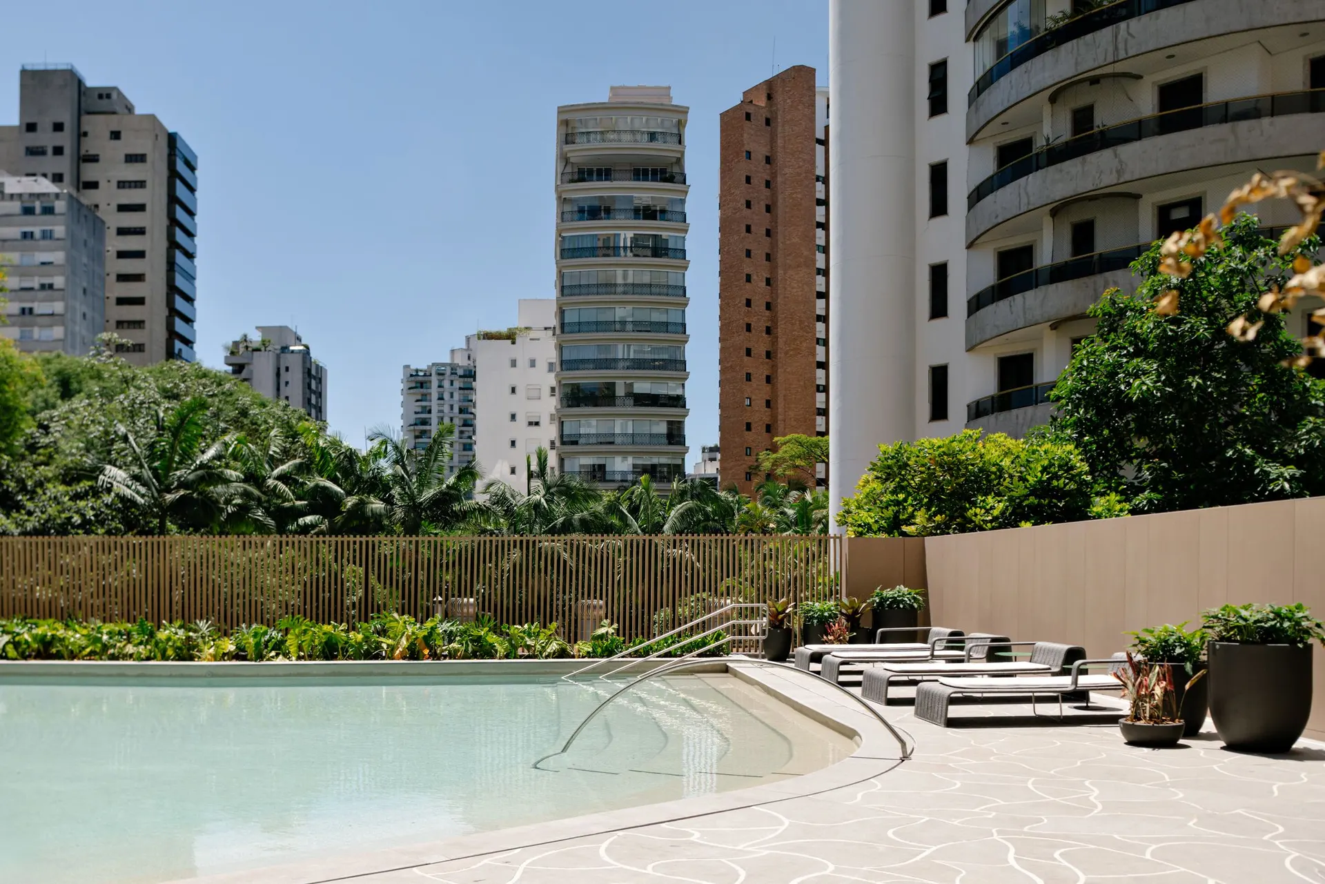 A piscina descoberta de desenho curvo, com o deck, as espreguiçadeiras e o jardim, entre os prédios do bairro.
