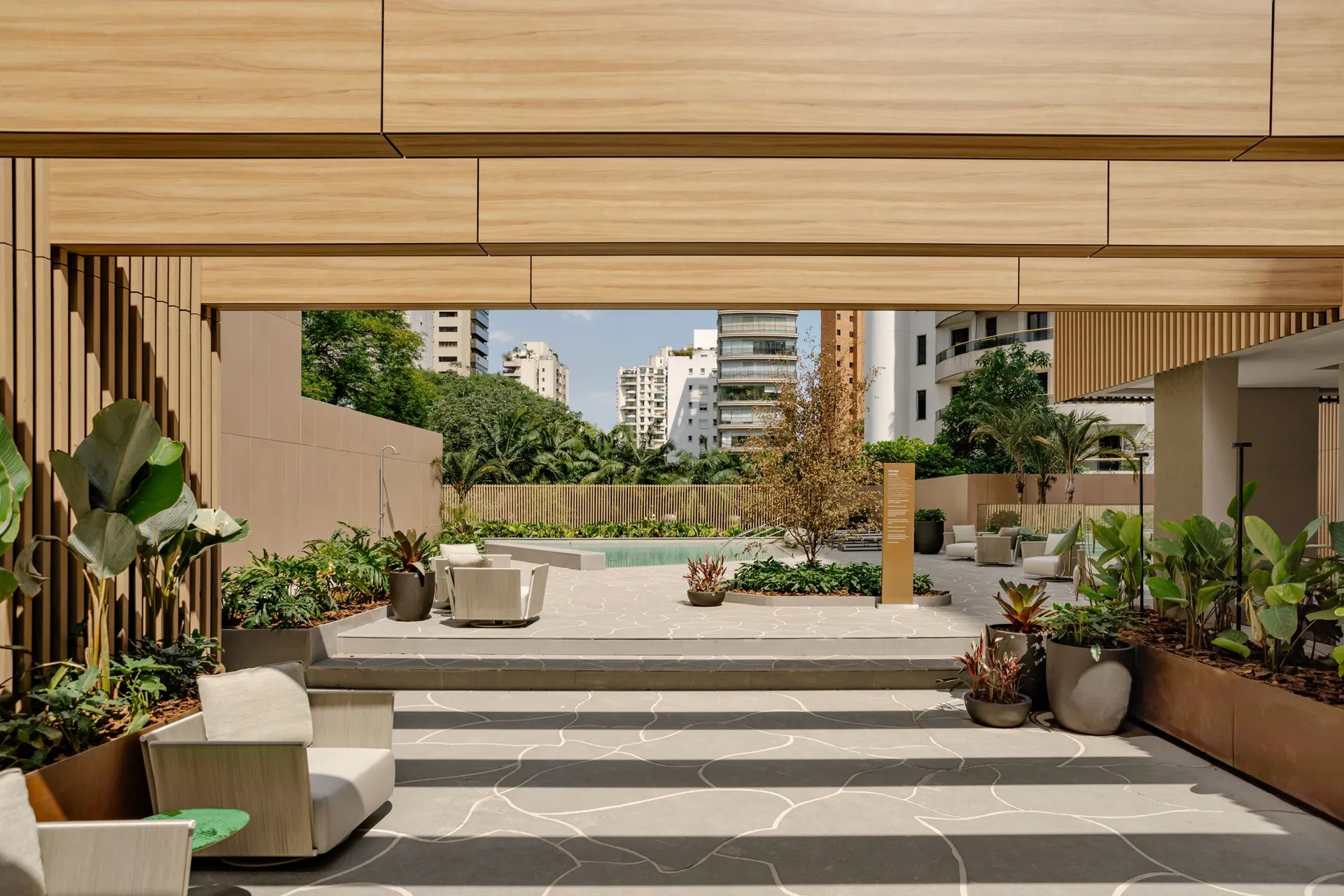 A praça externa sob o pergolado de madeira, com poltronas, vasos e a piscina ao fundo.
