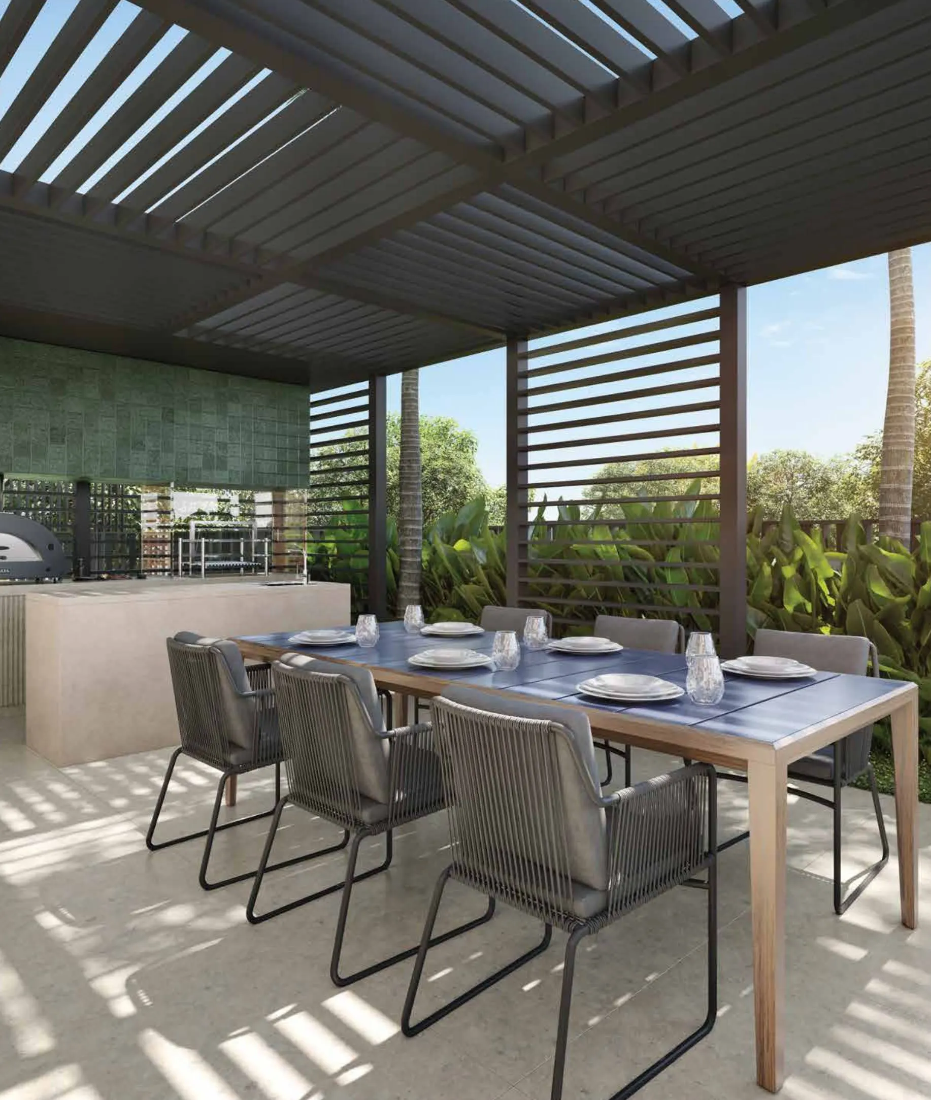 A churrasqueira sob uma pérgola de lâminas, com mesa comprida e cadeiras ao sol.