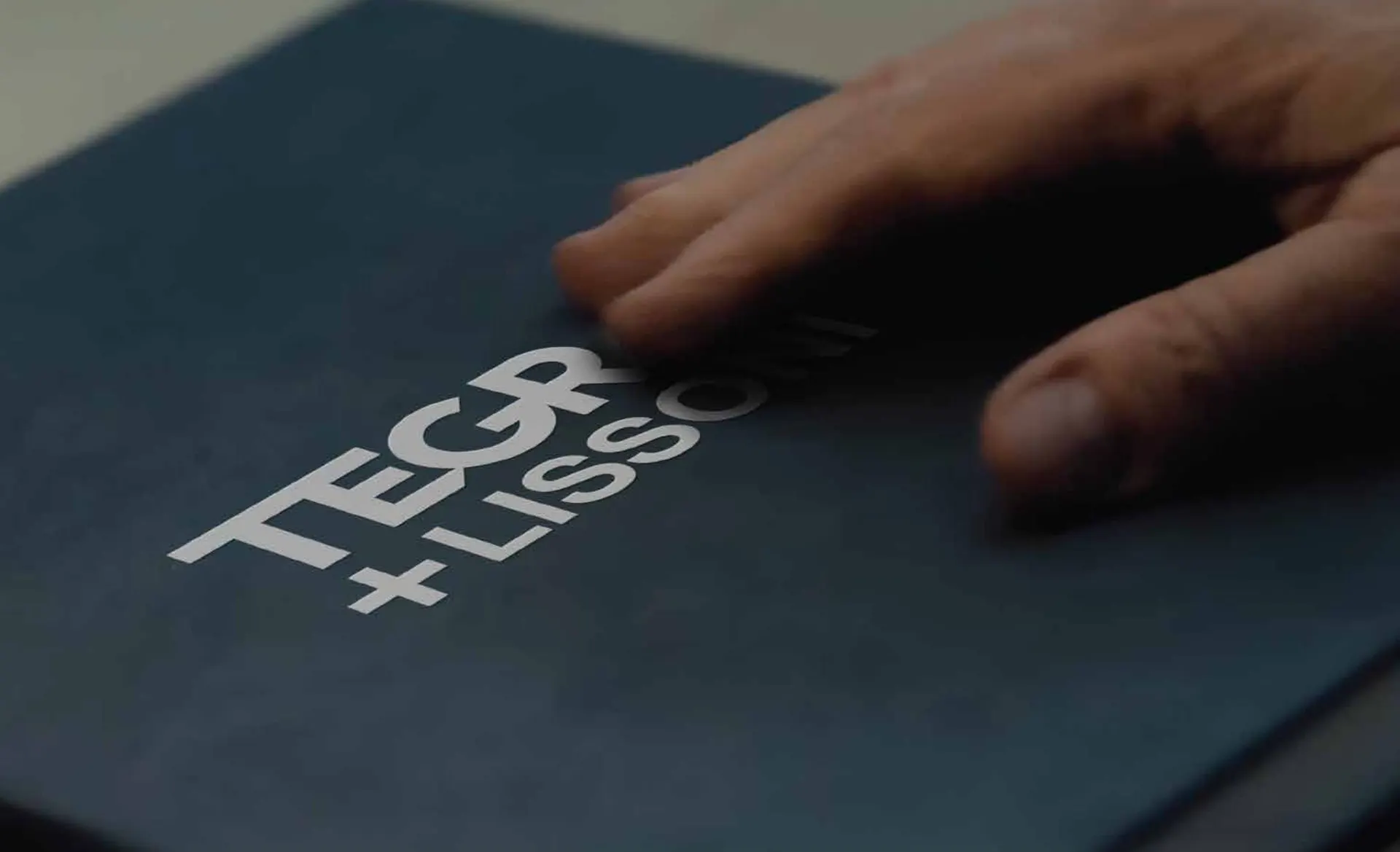 Uma mão sobre a capa petróleo do book, com TEGRA + LISSONI gravado em relevo.