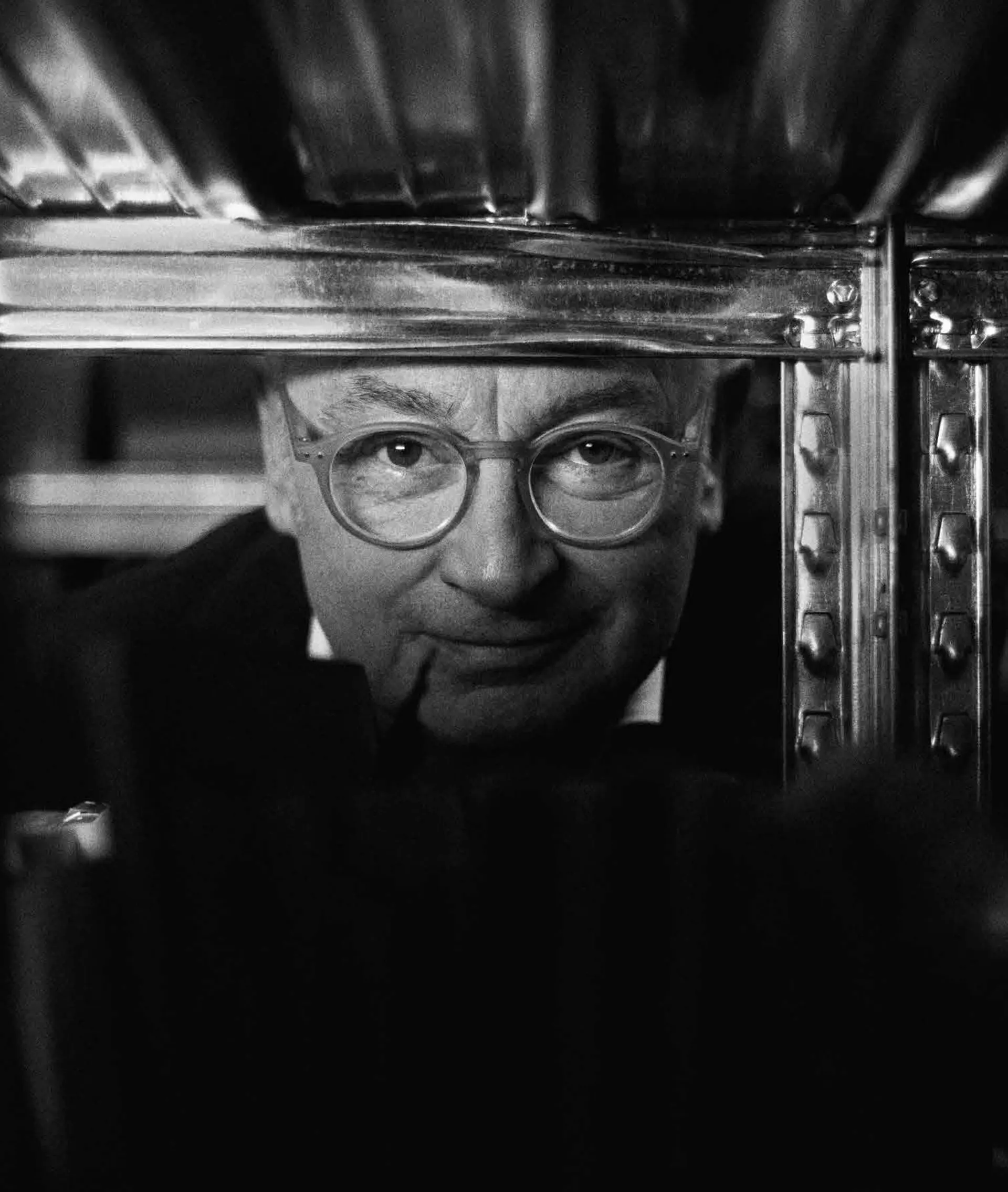 Retrato em preto e branco de Piero Lissoni, de óculos redondos, entre as prateleiras de uma estante de metal.