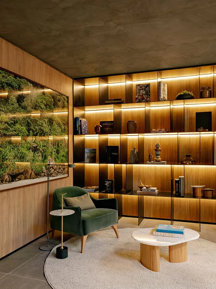 Uma sala das áreas comuns com a estante de madeira iluminada, a poltrona verde, a mesa redonda e um jardim vertical atrás do vidro.