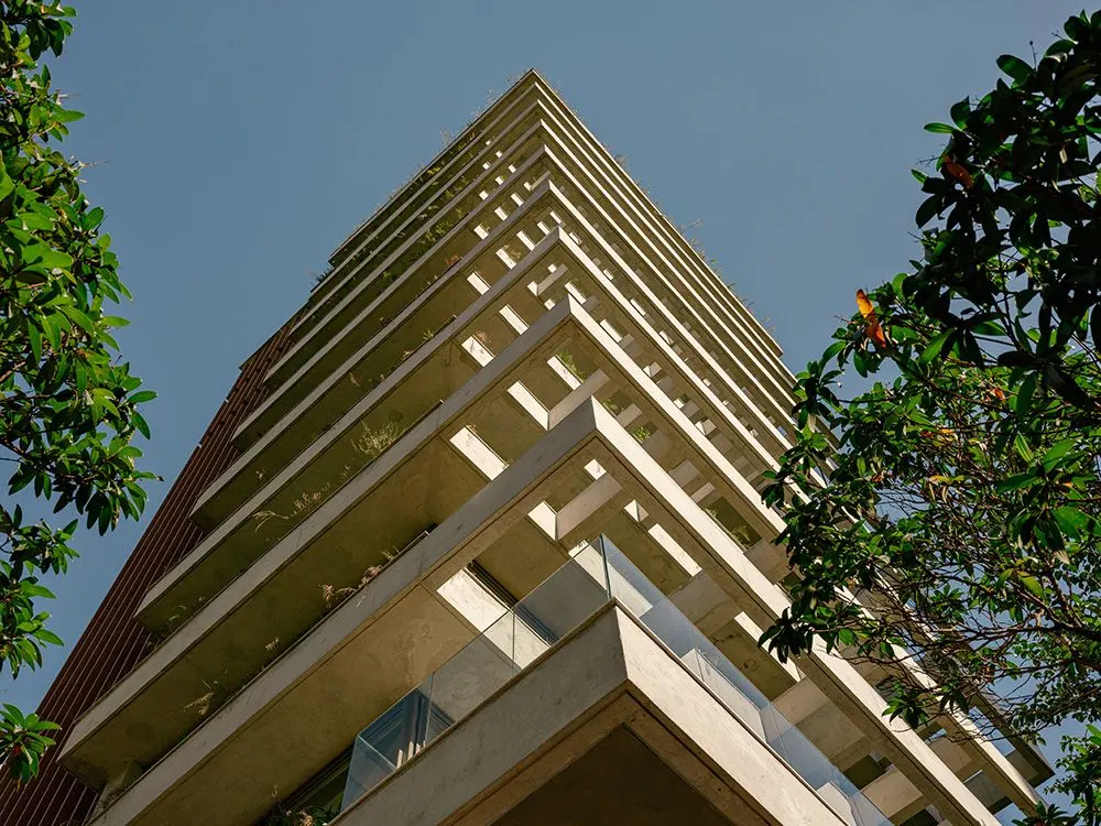 A esquina da torre vista de baixo: os quadros de concreto em balanço, andar sobre andar, com plantas nas jardineiras e uma árvore à direita.