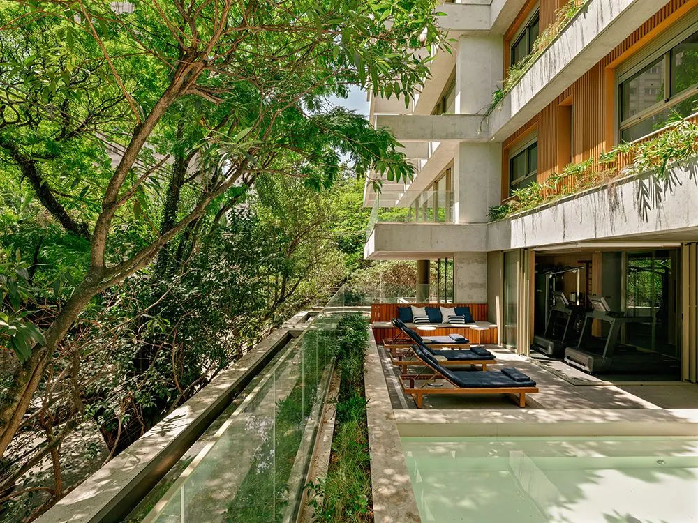 A raia da piscina ao lado do jardim, vista do alto, com as espreguiçadeiras, o sofá e a academia de vidro ao fundo, sob os andares da torre.
