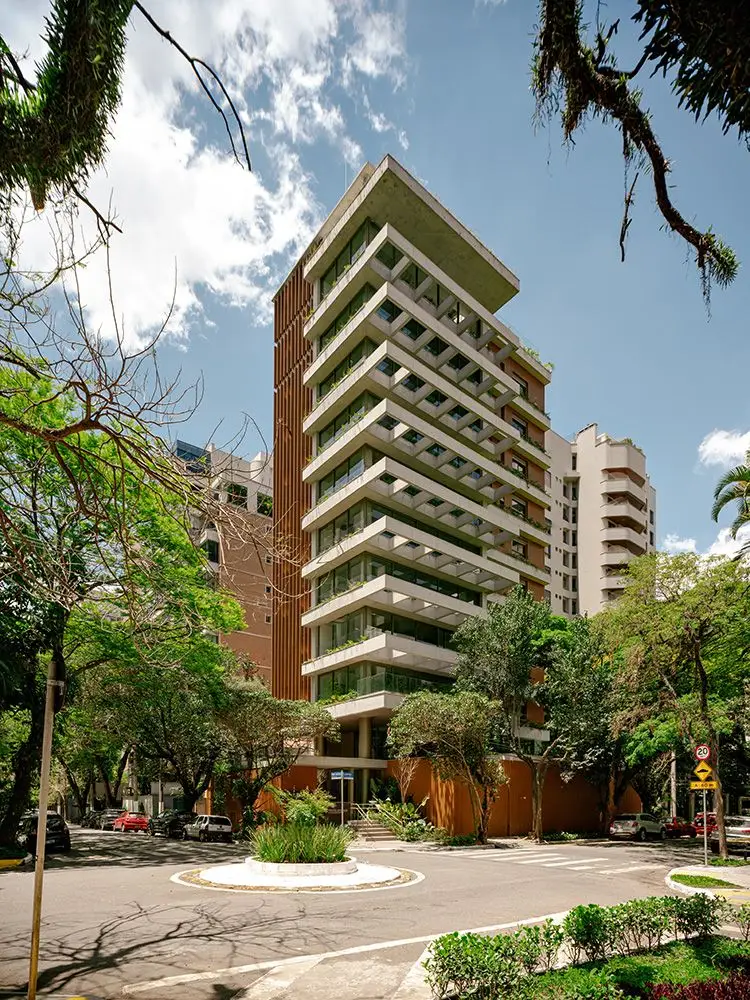 A torre da Casa Pereira Coutinho pronta, vista da rua: os quadros de concreto empilhados, com plantas em cada andar, entre as copas das árvores da calçada e o céu azul.