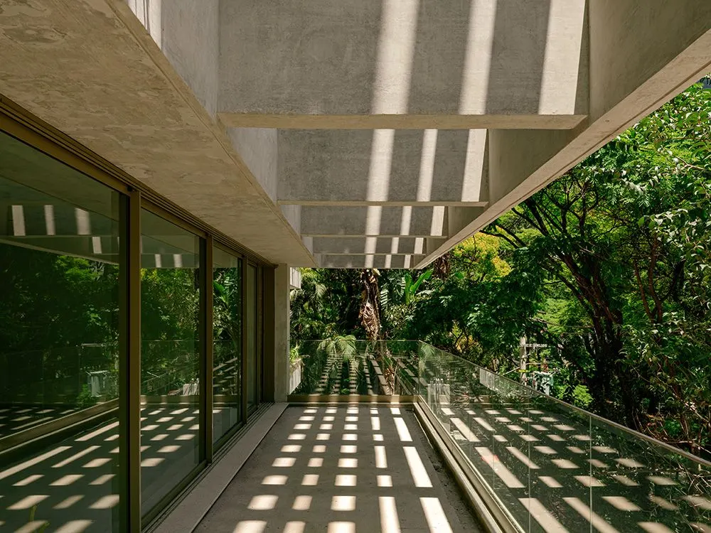 A varanda sob o quadro de concreto: o vidro do apartamento à esquerda, o guarda-corpo de vidro à direita, as árvores em volta e a sombra em grade das vigas no piso.