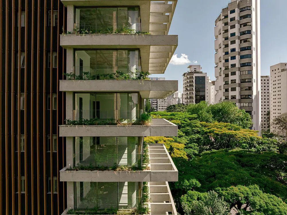As varandas da torre com plantas nas jardineiras, de perto, e os prédios da cidade ao fundo, sobre as árvores.