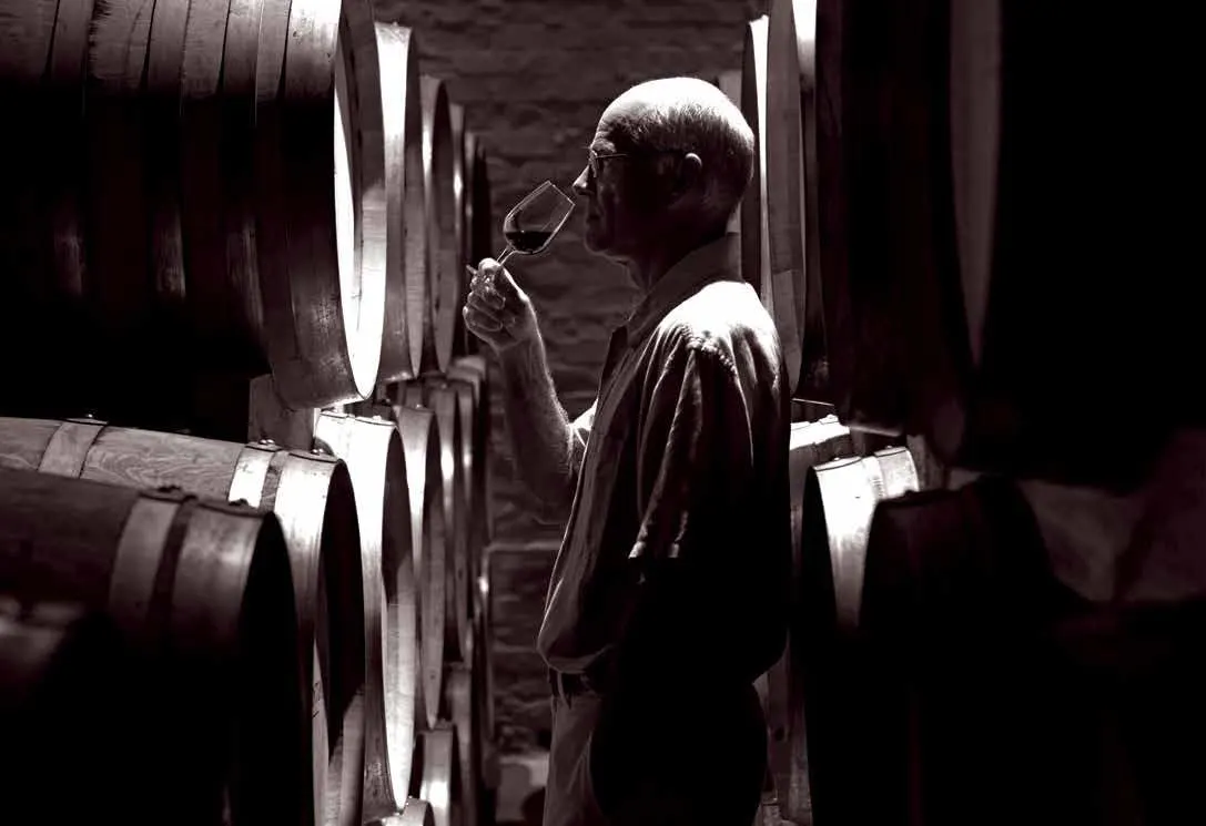 Um homem de cabelos brancos prova vinho numa adega escura, entre fileiras de barris de carvalho, em preto e branco.