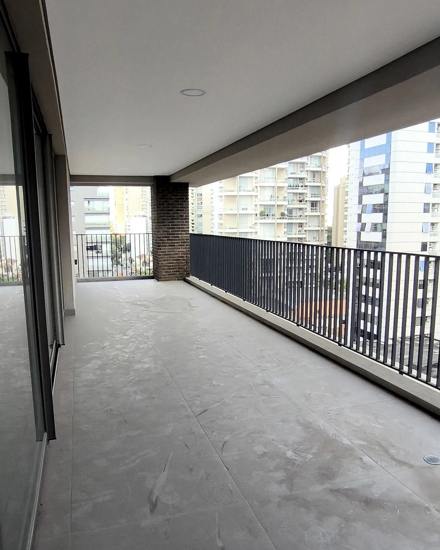 O terraço da unidade 102 no contrapiso: o piso cinza, o teto liso, o guarda-corpo de barras escuras e os prédios do bairro no fim.