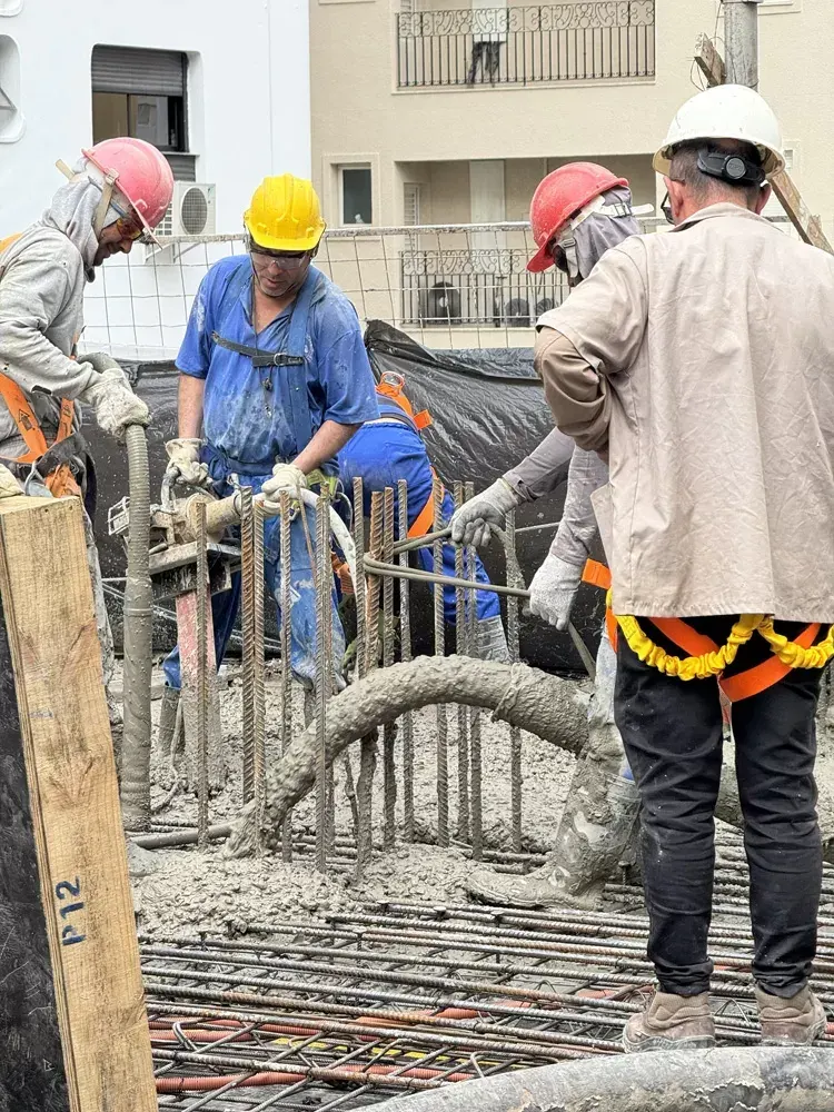 Operários de capacete e colete concretam a laje: a mangueira de concreto, as armaduras no chão e as escoras em volta.