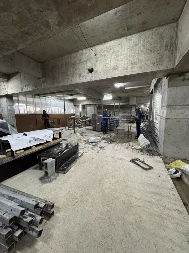 Um subsolo em obra: a laje de concreto aparente com luminárias provisórias, perfis metálicos no chão e a rampa ao fundo.