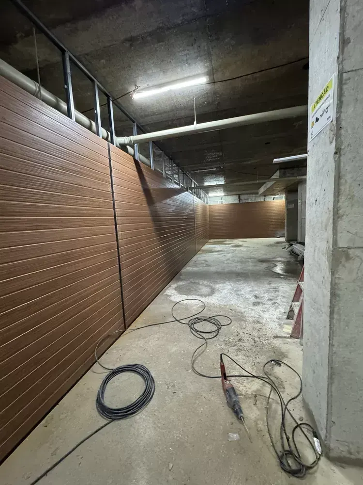 Um corredor do subsolo com a parede revestida de madeira escura, a laje de concreto e cabos de obra no chão.