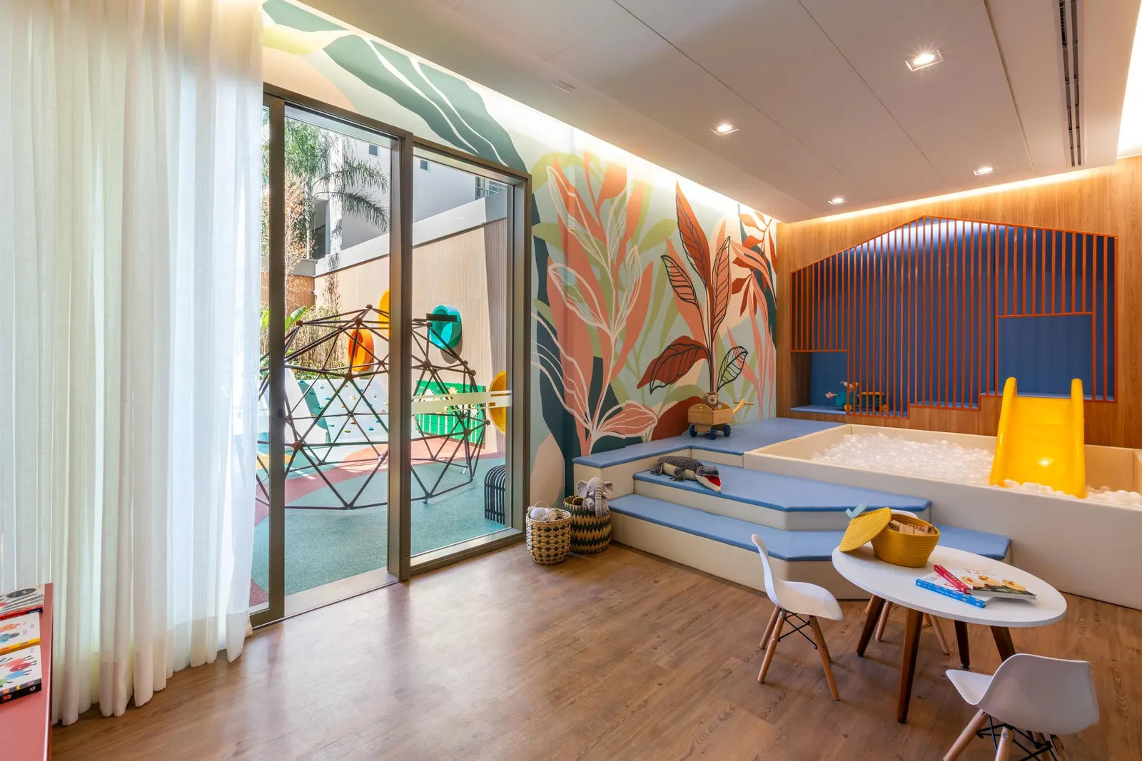 A brinquedoteca: piscina de bolinhas, escorregador amarelo, mesinha com cadeiras e um painel de folhagens na parede.