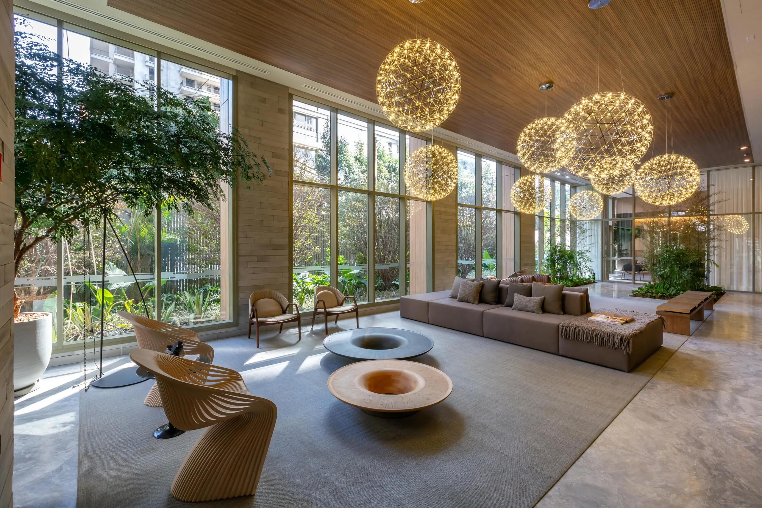 O lobby de pé-direito alto: luminárias esféricas de luz acesa penduradas do forro de madeira, sofás e poltronas sobre o tapete e a parede inteira de vidro aberta para o jardim.