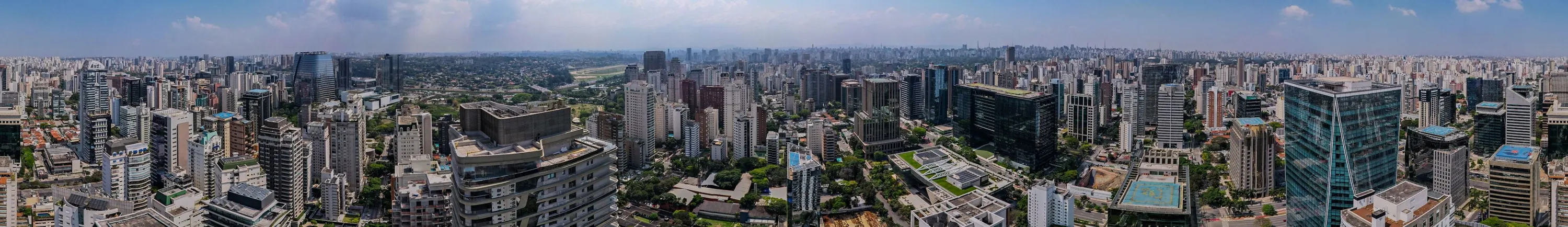 Panorama de 360 graus do Itaim visto do alto, sobre o terreno: torres de escritórios, prédios residenciais, a Marginal Pinheiros, a pista do Jockey Club, o verde do Ibirapuera ao longe e o céu claro.