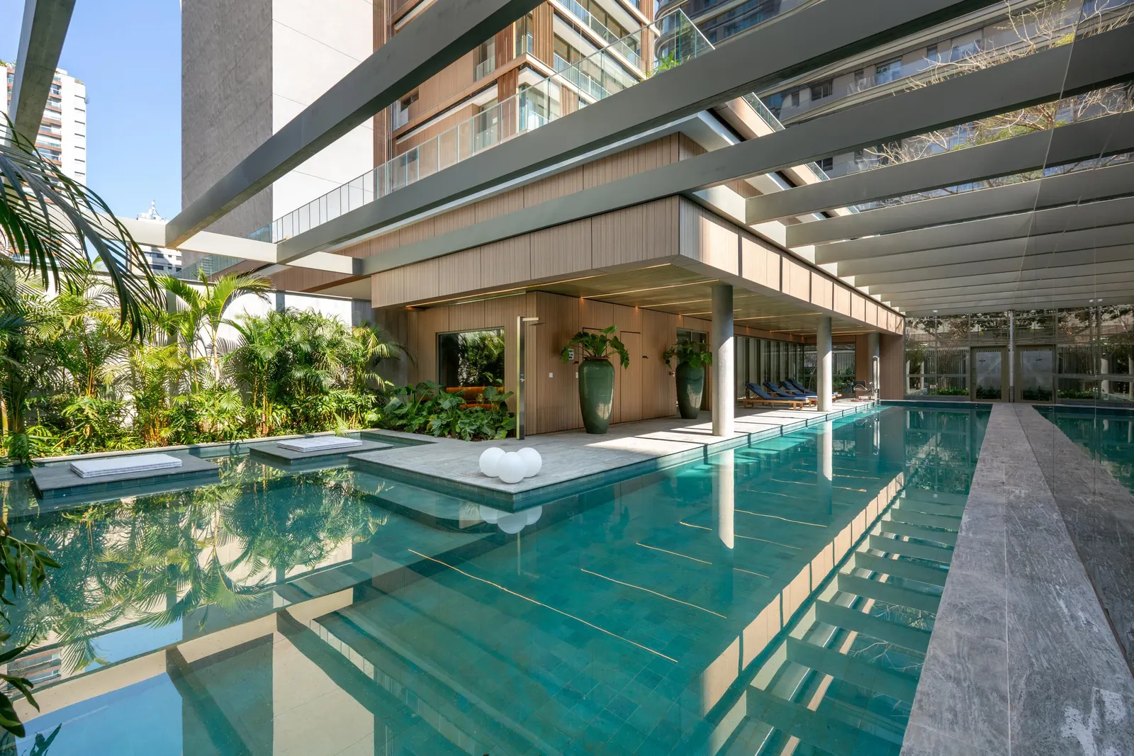 A piscina comprida sob a pérgola, com degraus na água, o deque de pedra e o jardim tropical em volta.